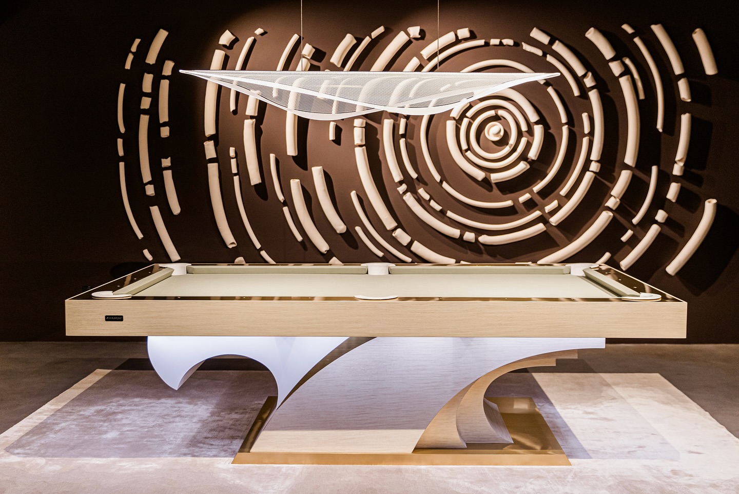A modern billiards table with a sculptural base stands on a light rug. Above it hangs a contemporary white light, and the wall behind features a large abstract circular art installation.