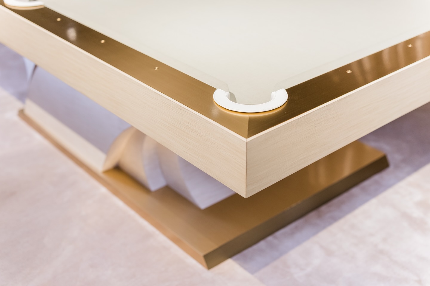 A close-up of a modern, light-colored billiards table with gold trim and a unique geometric base, focusing on the corner pocket and smooth surfaces.