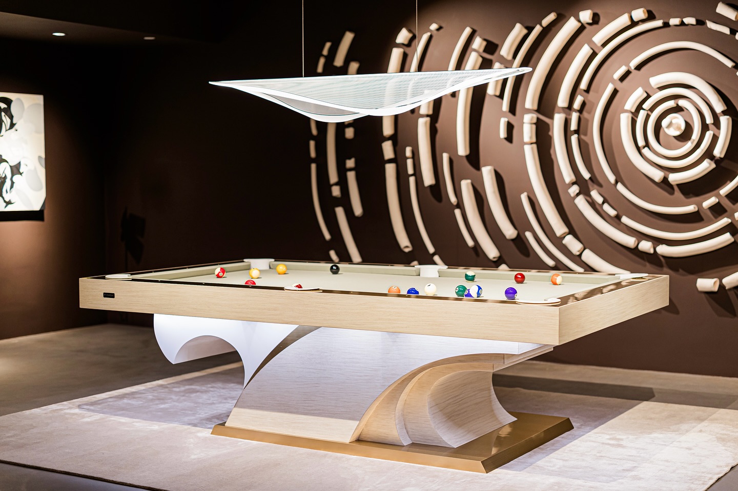 A modern billiards table with a sculptural wooden base stands on a light rug. Above it hangs a sleek, geometric light. The brown wall behind features abstract circular art with raised, curved shapes.