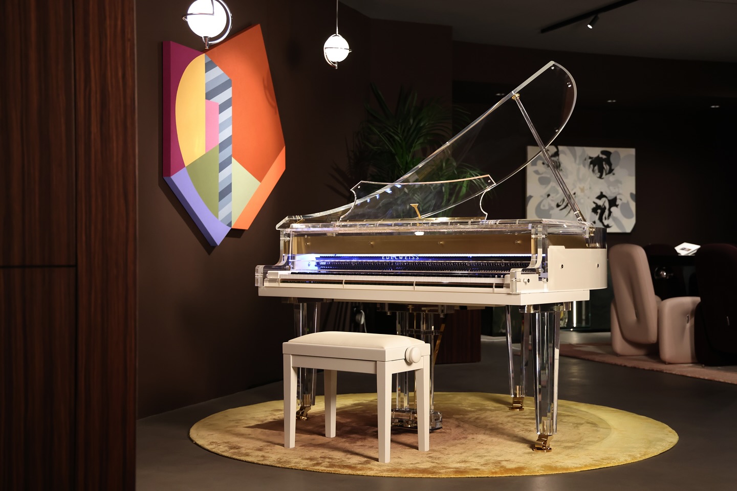 A transparent grand piano with a white bench sits on a round yellow rug in a stylish room, featuring modern art on the brown walls and elegant lighting overhead.