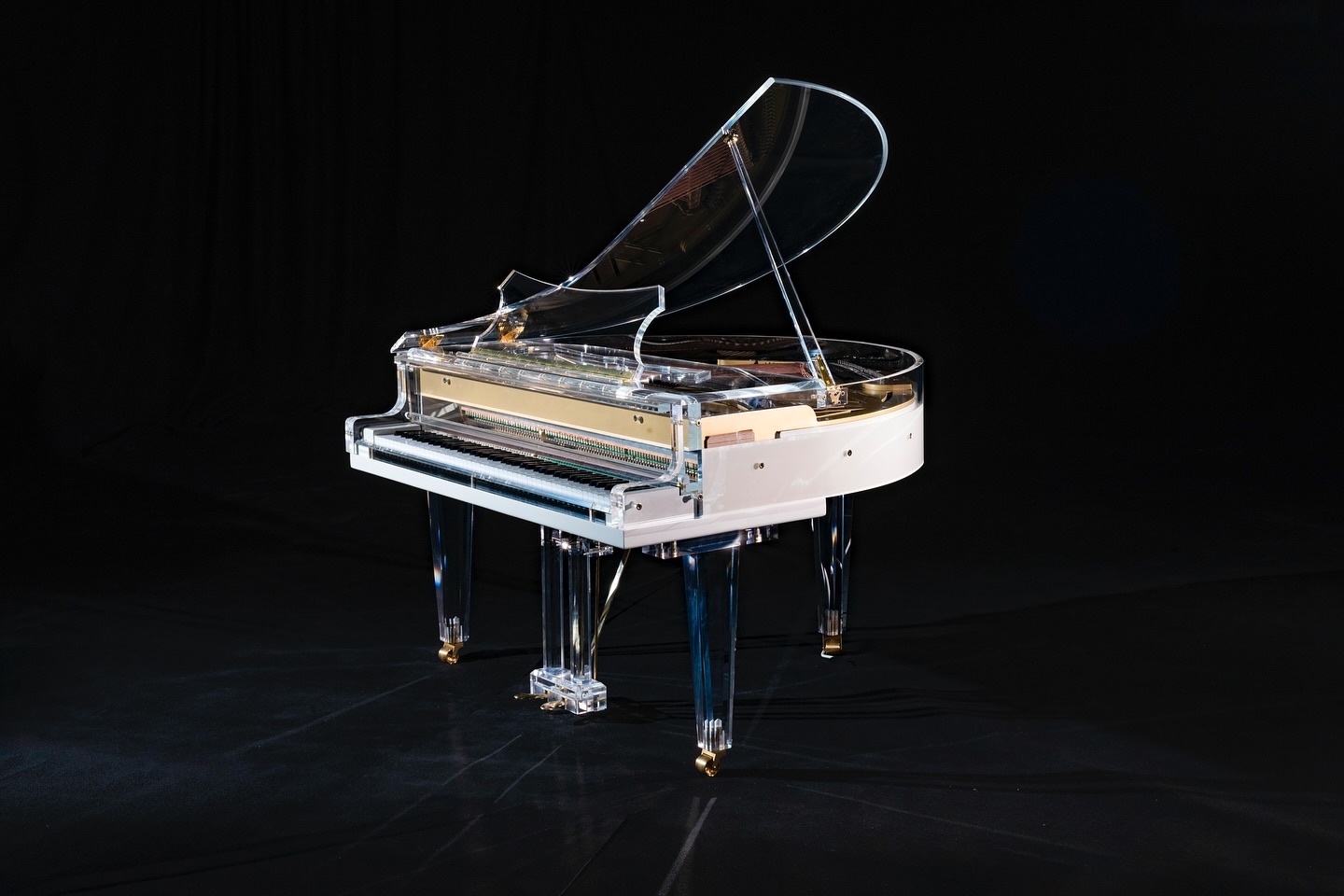 A transparent grand piano with a clear acrylic body, lid, and legs, accented with gold hardware, is displayed against a black background.