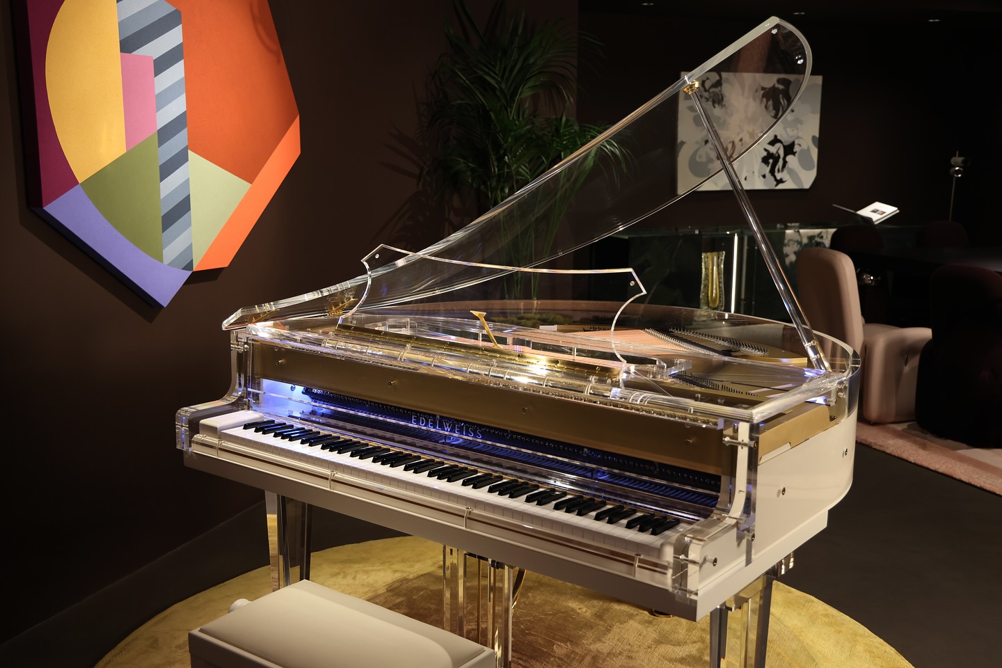 A transparent grand piano with a white base is displayed on a round yellow rug in a stylish room, decorated with modern art and plants.