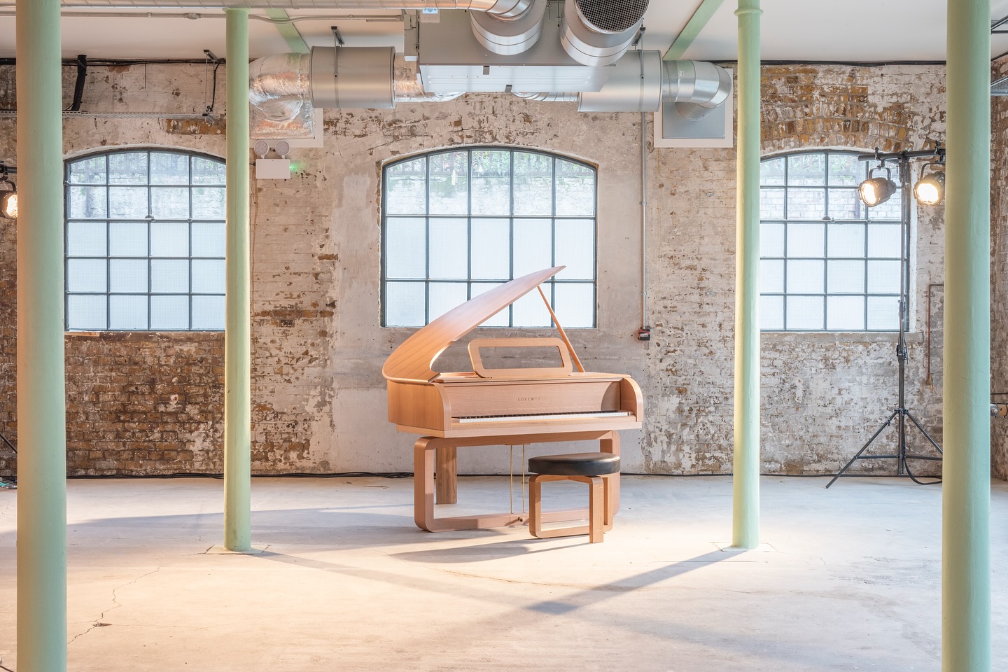 A modern wooden grand piano with a matching bench is placed in an industrial-style room with exposed brick walls, large windows, and overhead ductwork. Soft natural light fills the space.