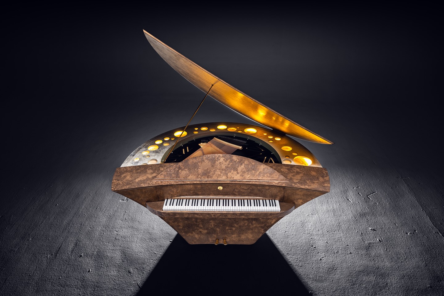 A unique, futuristic grand piano with a metallic, weathered finish, illuminated circular accents around the open lid, and a spotlight highlighting the piano against a dark background.