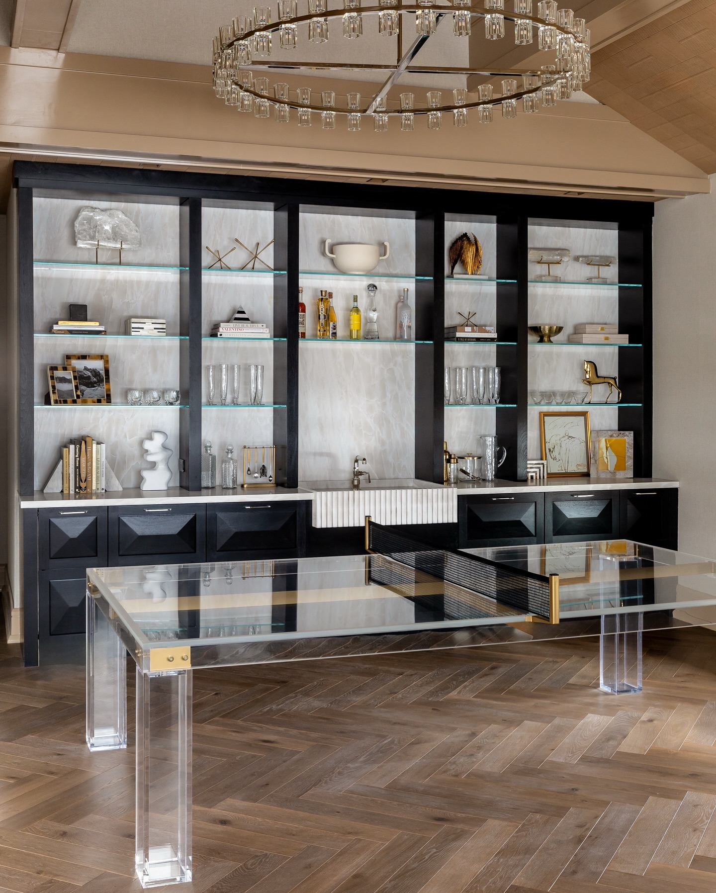 A modern room features a glass ping pong table with clear legs, a herringbone wood floor, and black built-in shelves displaying decorative items, books, and glassware. A round chandelier hangs above.