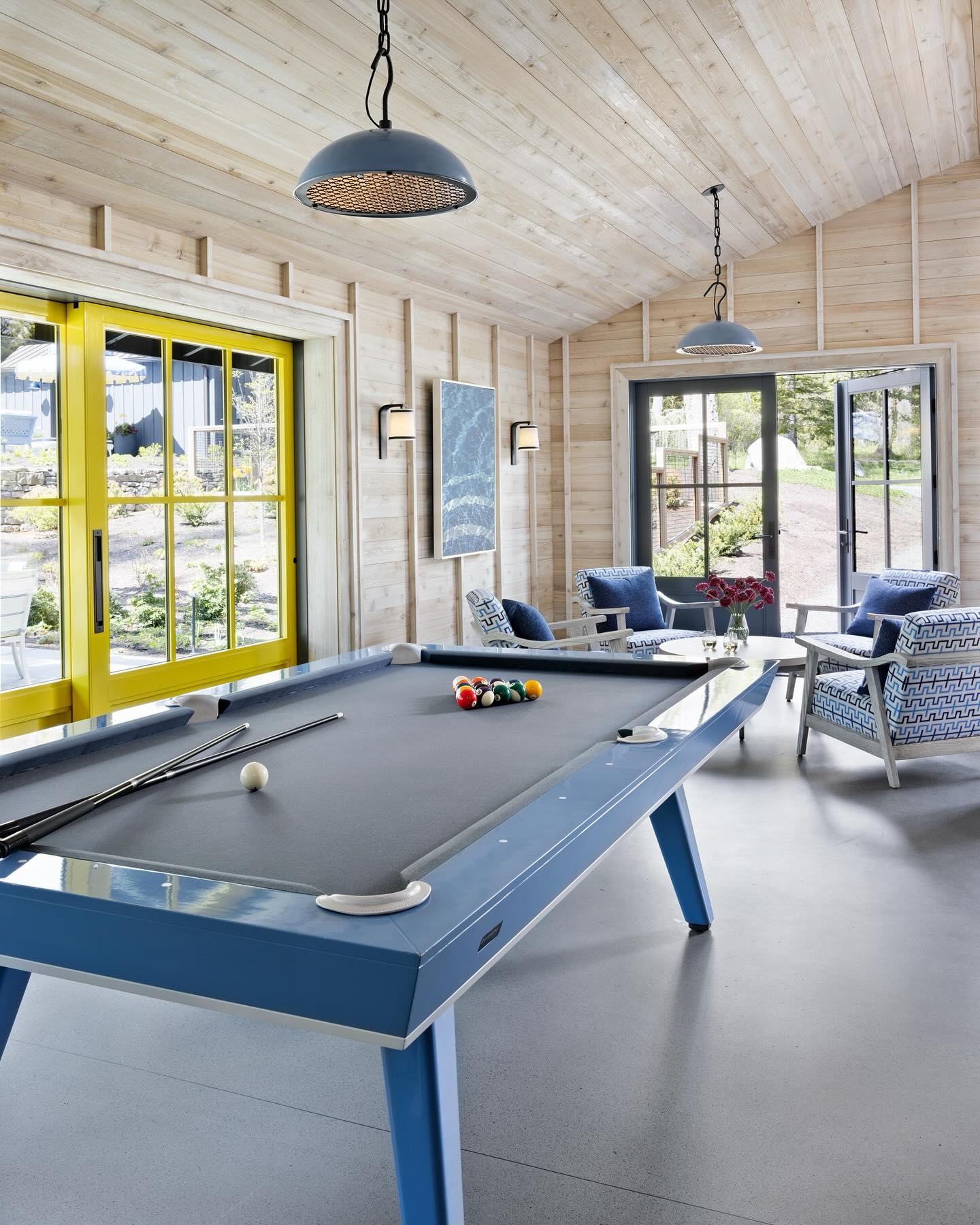 A modern game room with light wood walls, yellow sliding doors, large windows, a blue pool table, and cozy seating with decorative cushions. Pendant lights hang from the ceiling.