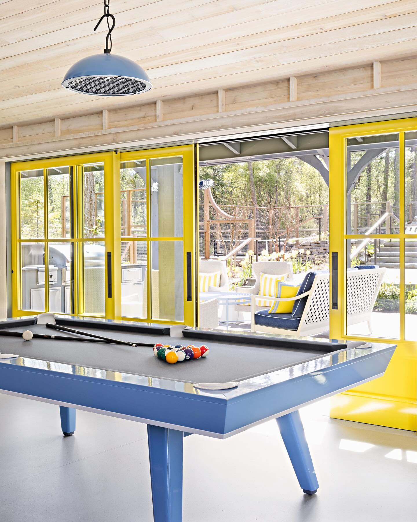 A modern game room with a blue pool table, billiard balls, and cues. Large yellow-framed glass doors open to a sunny patio with white furniture and yellow-striped cushions. Light wood ceiling and bright, airy atmosphere.