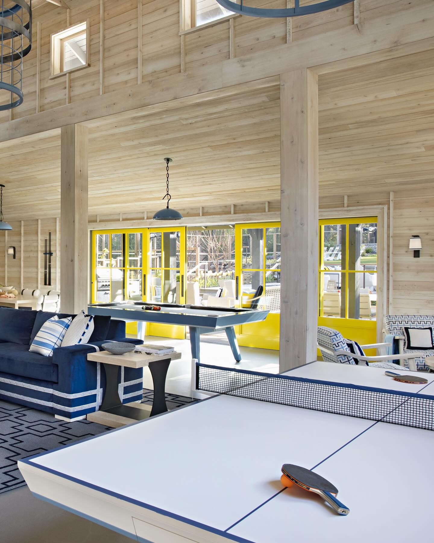 A bright, modern game room with wood-paneled walls, a navy sofa, ping pong table, and foosball table. Large yellow-trimmed windows let in natural light, creating an inviting space for relaxation and play.