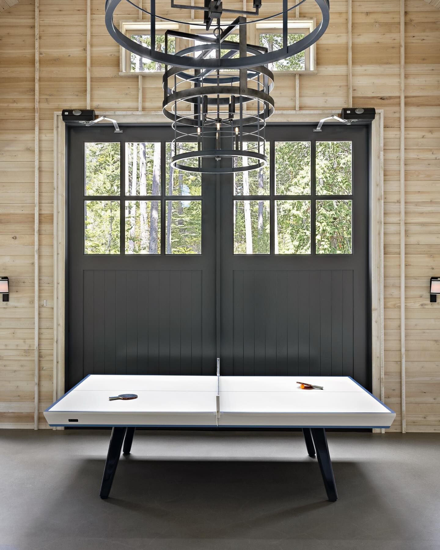 A modern white ping pong table with two paddles stands in a wooden room, in front of large, dark double doors with windows. Industrial-style light fixtures hang from the ceiling. Trees are visible through the windows.