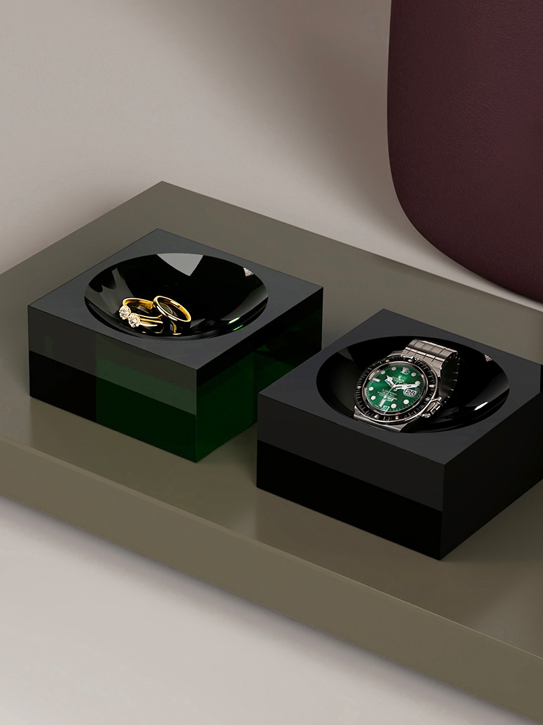 Two black, square trays on a gray surface hold jewelry; the left tray contains gold rings, and the right tray holds a silver wristwatch with a green face.