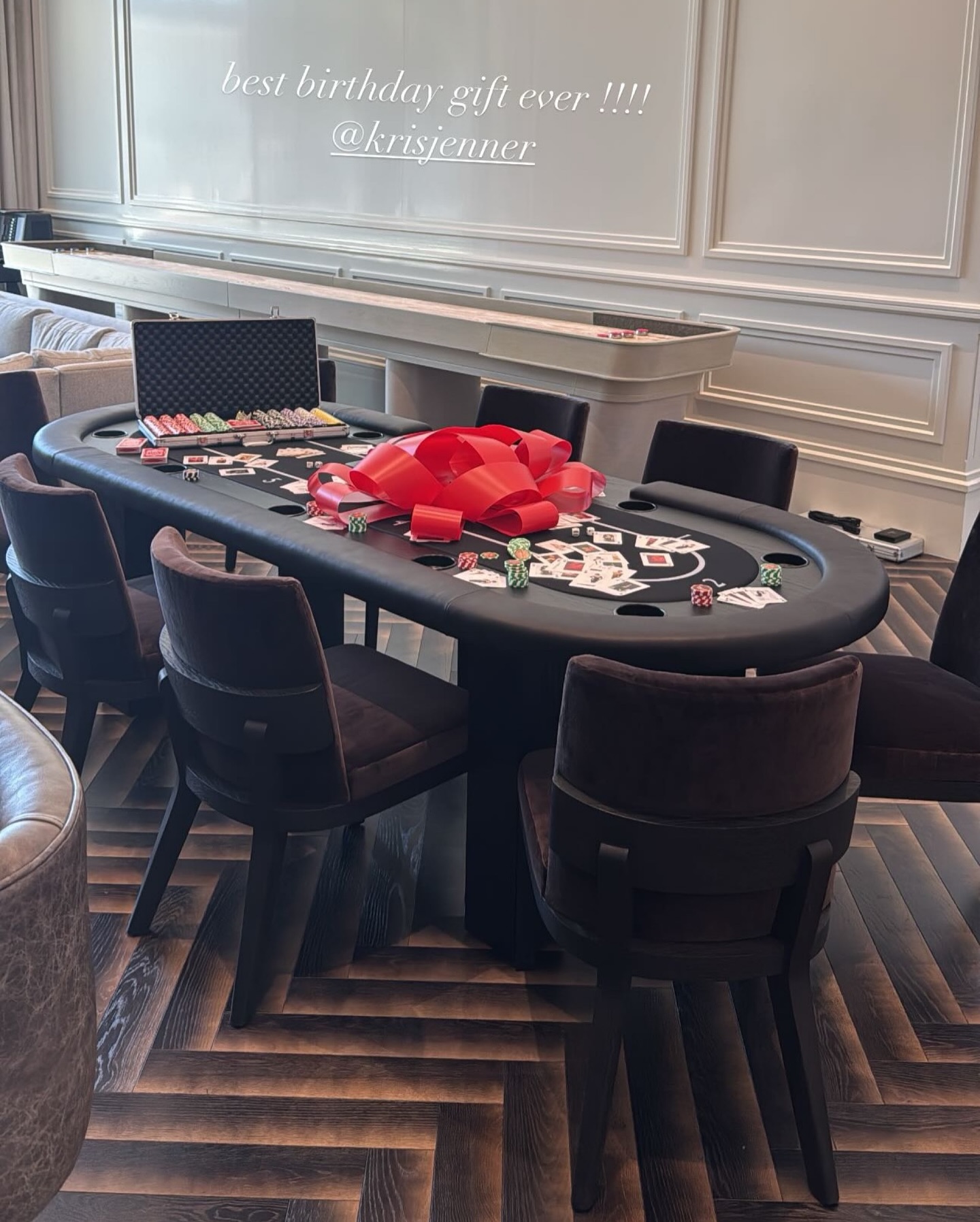 A poker table with chairs is set up in a stylish room. Poker chips, cards, and a case are arranged on the table, which is topped with a large red gift bow. Text above reads, best birthday gift ever !!!! @krisjenner.