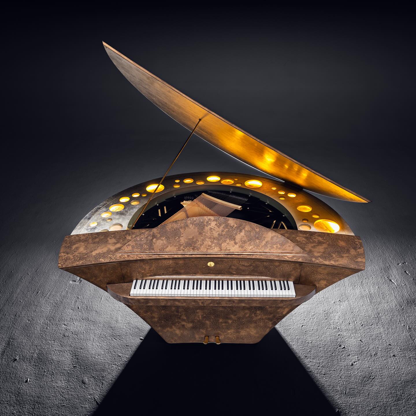 A modern grand piano with a unique, futuristic design, featuring a circular pattern of golden lights around the strings and a metallic, angular lid partially open, viewed from above on a dark background.