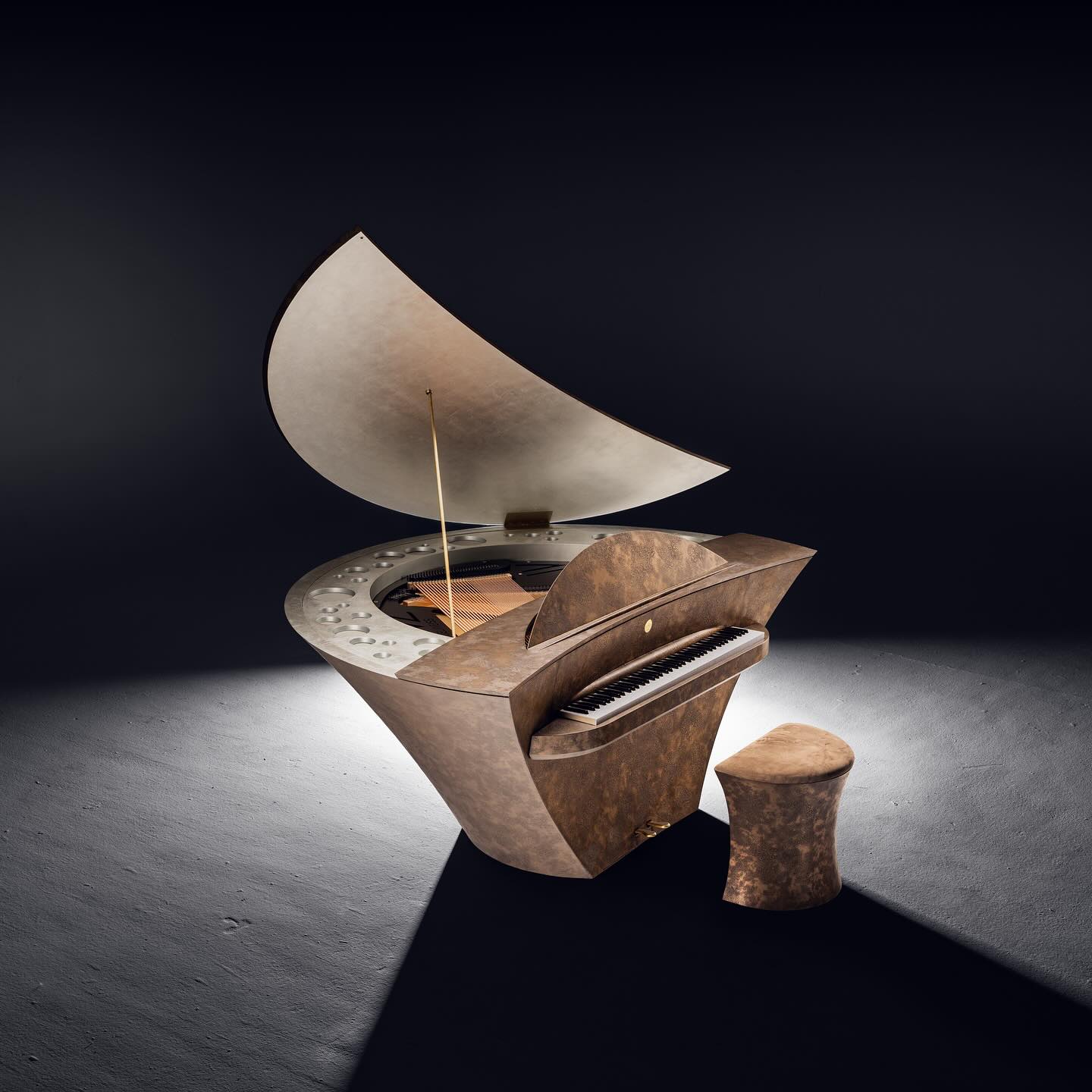 A modern, sculptural grand piano with a curved, asymmetrical design and matching bench, set under focused lighting on a dark floor. The piano lid is partially open, revealing the strings inside.