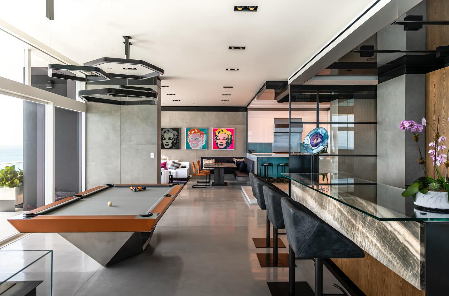 Modern living space with a pool table, bar area with stools, glass decor, and pop art portraits on the wall. Large windows let in natural light and offer an ocean view; orchids and plants decorate the room.