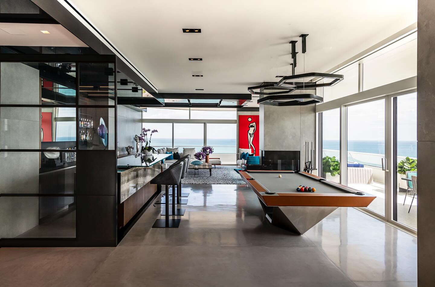 Modern open-concept living space with a sleek pool table, bar area, large windows offering ocean views, contemporary furnishings, and coastal decor.