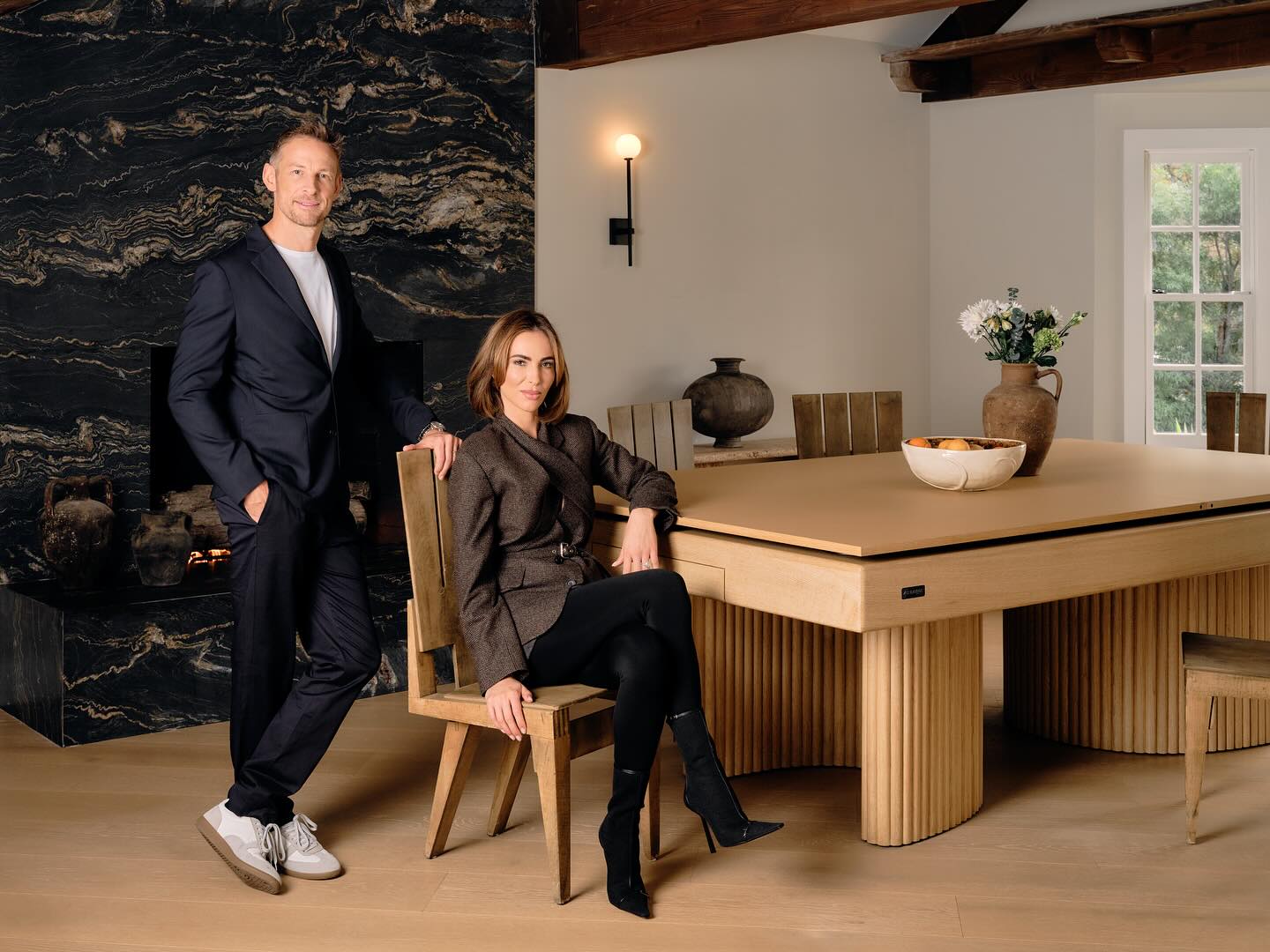 A man in a dark suit stands beside a woman seated at a modern wooden dining table—designed by an interior designer—in a stylish room with a black marble fireplace, rustic decor, and large windows letting in natural light.