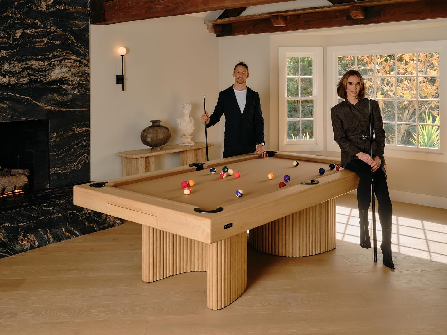 A man stands at one end of a bespoke game table holding a cue stick, while a woman in a blazer sits on its edge. The room, styled by an interior designer, features a black marble fireplace, wooden beams, and a large window.