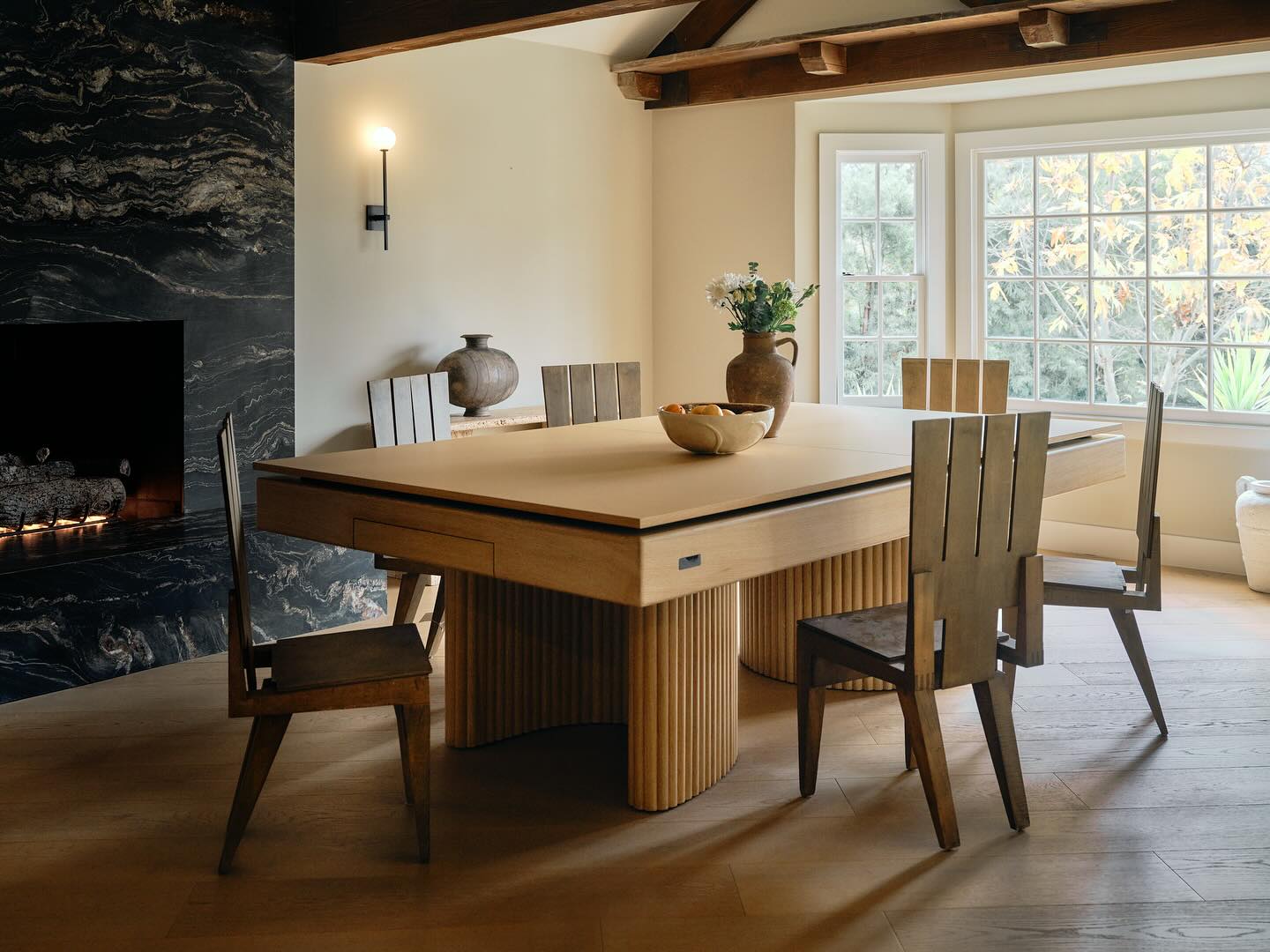 A modern dining room with a bespoke convertible game table and four matching chairs sits near a large window. Decorative vases and a bowl of fruit adorn the table, while a black marble fireplace and exposed wood beams add interior designer flair.