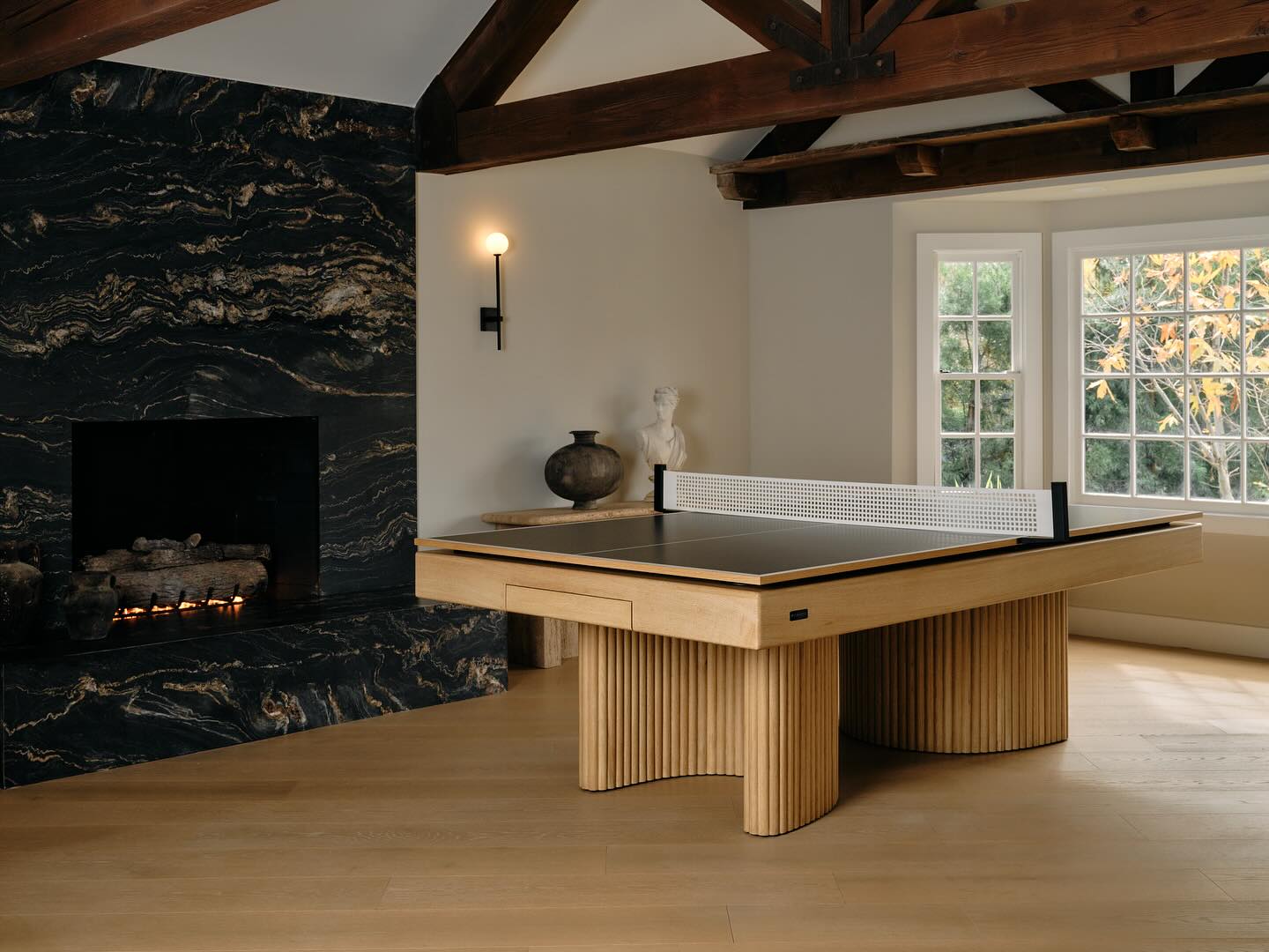 A modern room styled by an interior designer with exposed wooden beams, a black marble fireplace, a light wood ping pong table, and a window framing autumn trees. A statue and vase sit on the hearth, evoking Jenson Button’s love for refined spaces.