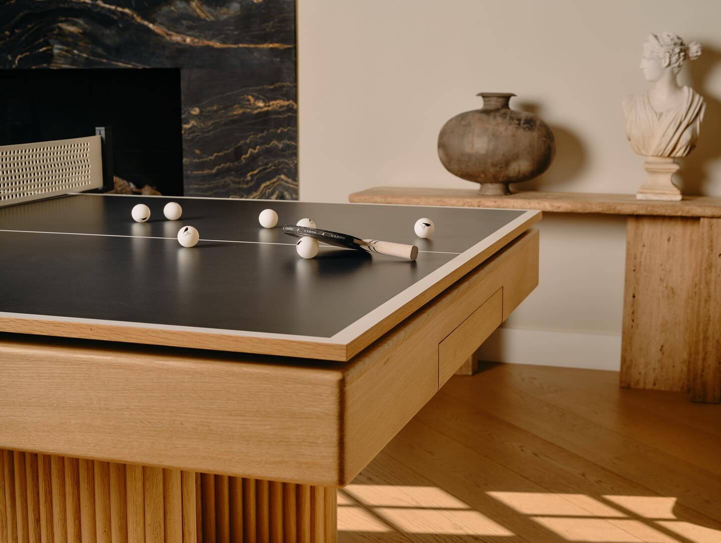 A modern ping pong table doubles as a bespoke convertible game table, set in a stylish room with wooden furniture, a decorative vase, marble fireplace, and classical-style bust on a console.