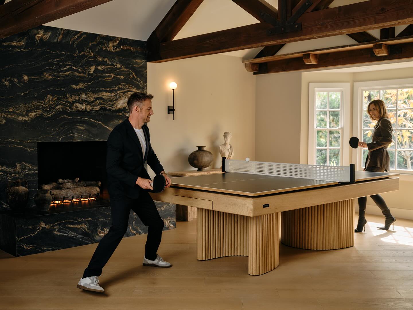 Two people play ping pong on a bespoke game table in a stylish living room with a fireplace, wooden beams, and large windows. One person is ready to hit the ball, while the other stands near the window holding a paddle.