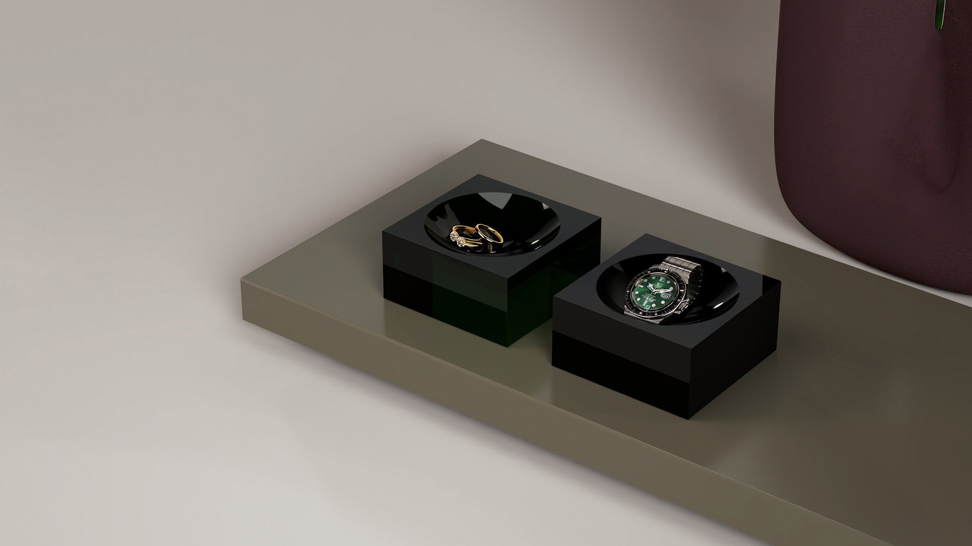 A gold ring and a green-faced wristwatch are displayed on separate black square trays atop a sleek, gray surface.