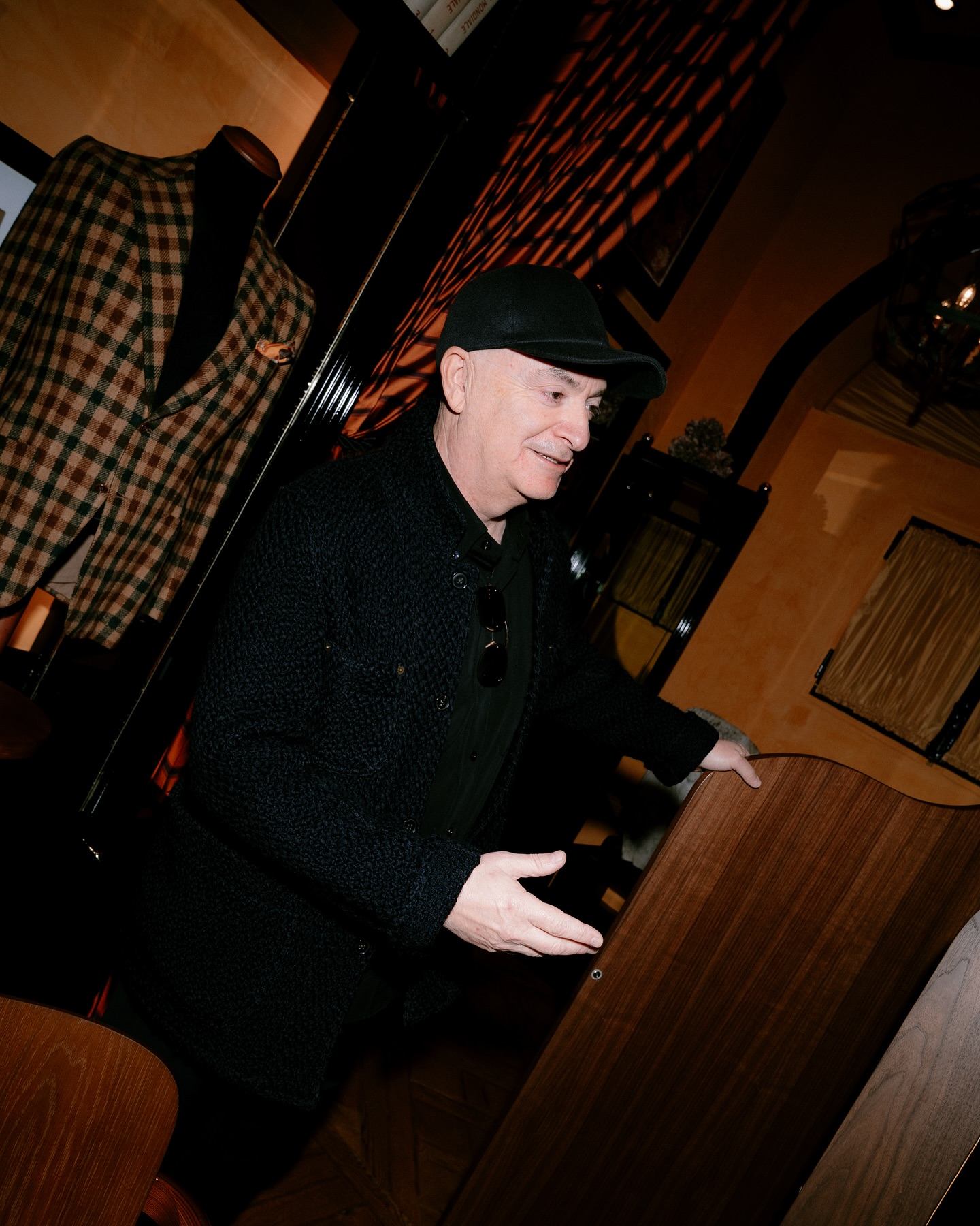 A man in a black cap and dark jacket stands indoors, gesturing with his hands near a wooden podium. Behind him is a mannequin wearing a plaid suit jacket and a warmly lit, stylish interior.