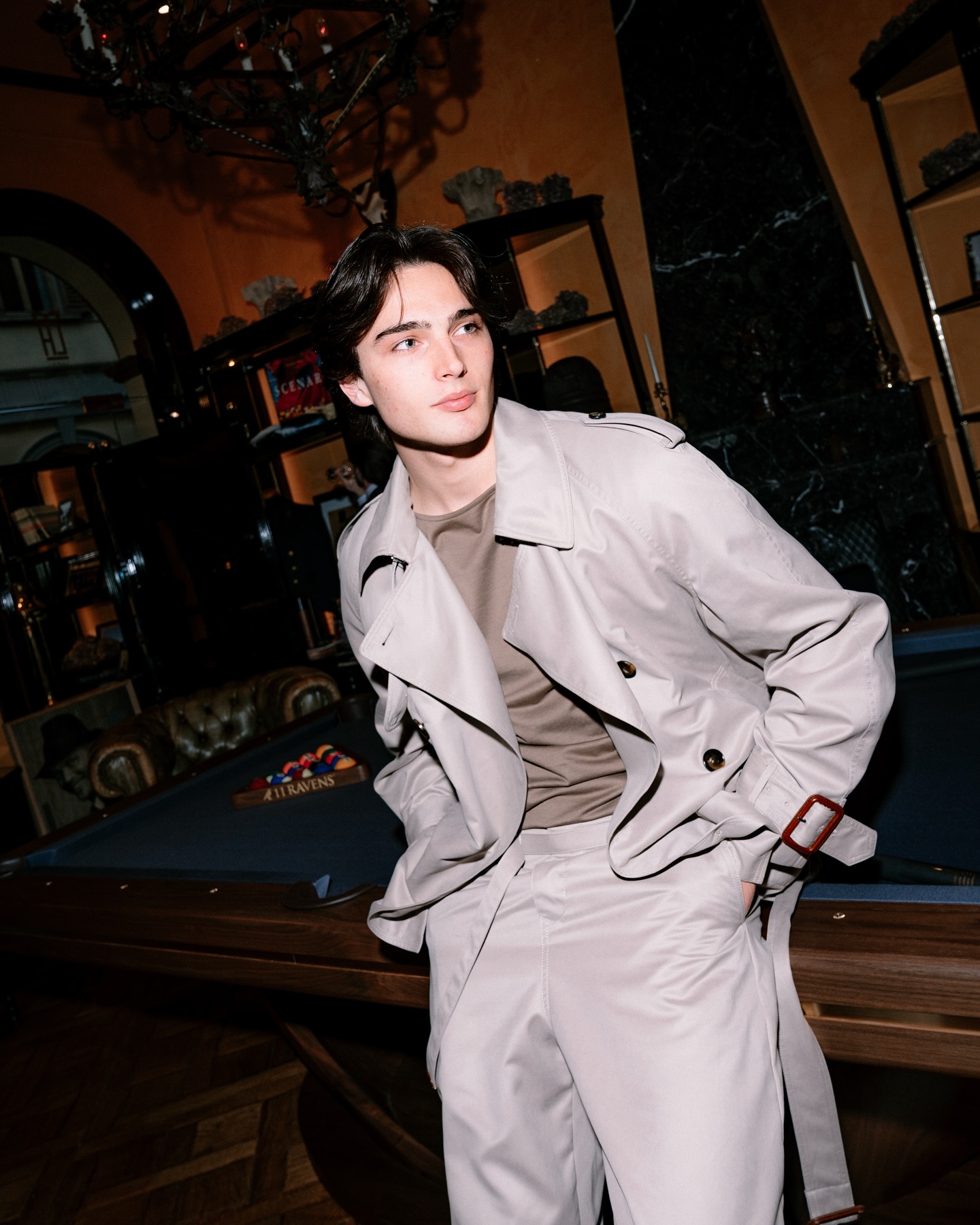 A young person with medium-length dark hair, wearing a light trench coat and matching pants, leans against a billiards table in a stylish, dimly lit room with warm-colored walls and vintage decor.