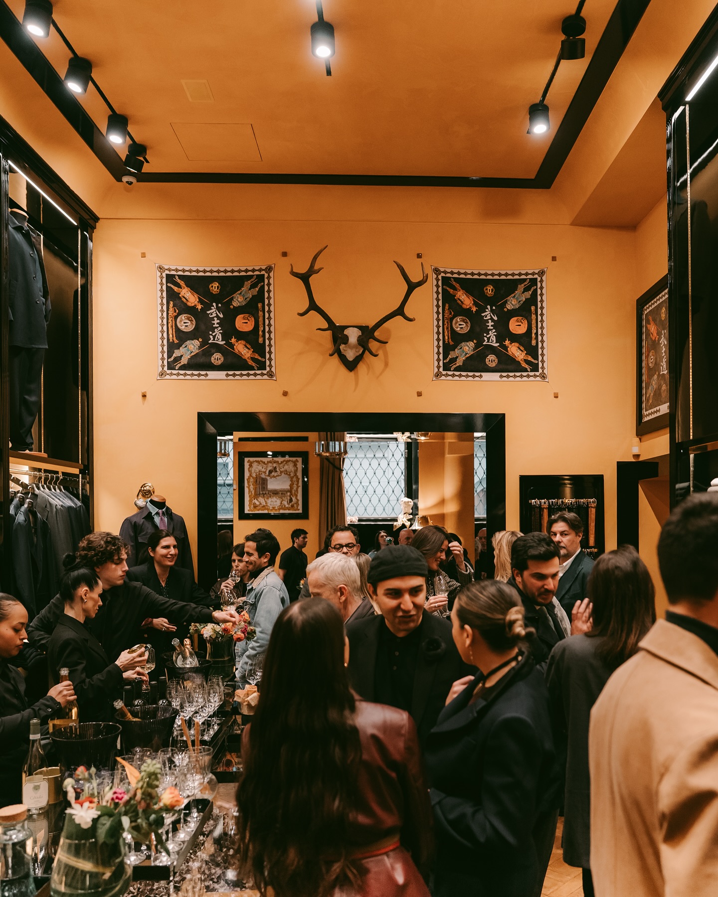 A stylish crowd mingles inside a warmly lit boutique with gold walls, dark shelves, and a deer antler mount above the entrance; racks of clothing and wine glasses fill the space.