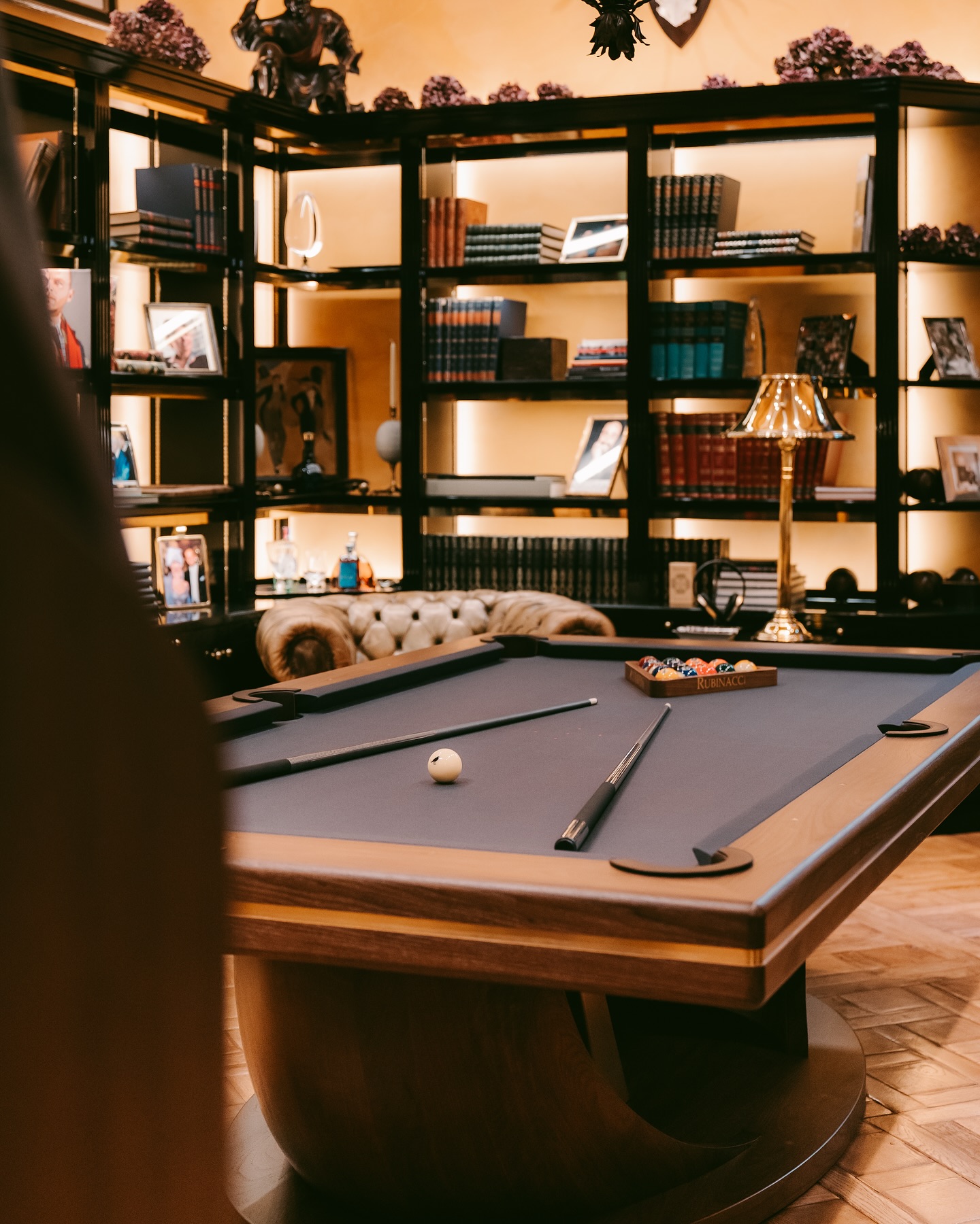A modern billiards table with cues and balls is centered in a stylish library room, featuring shelves filled with books, framed photos, and decorative items, with a sofa and a brass lamp in the background.