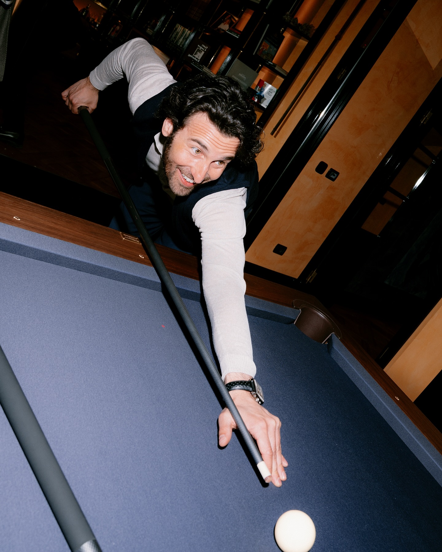 A man leans over a pool table, aiming a cue stick at the cue ball. He is smiling and focused, with shelves and warm lighting visible in the background.