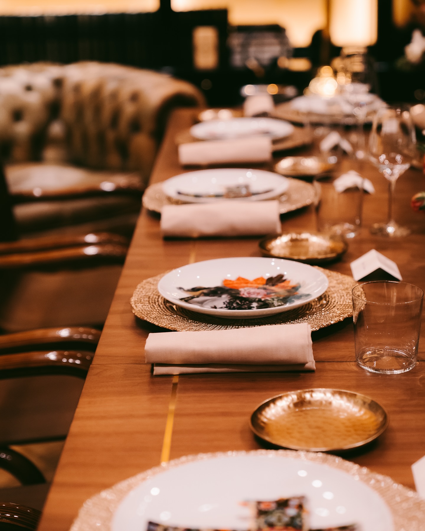 A wooden dining table set for a meal with woven placemats, plates, folded napkins, glassware, small gold dishes, and a plush chair in the background, creating an elegant ambiance.