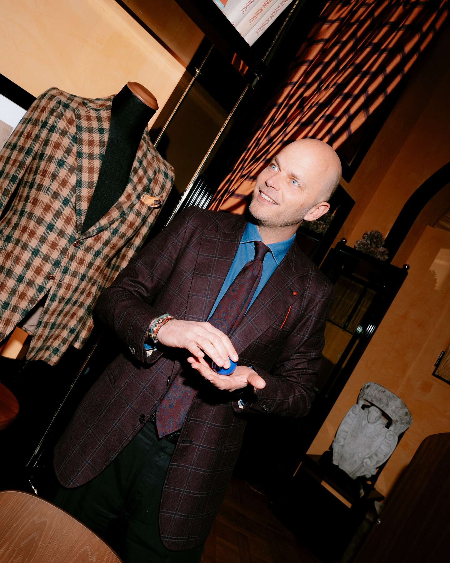 A smiling bald man in a plaid suit jacket and tie stands indoors beside a mannequin wearing a brown checked jacket, with warm lighting and patterned shadows on the wall behind him.