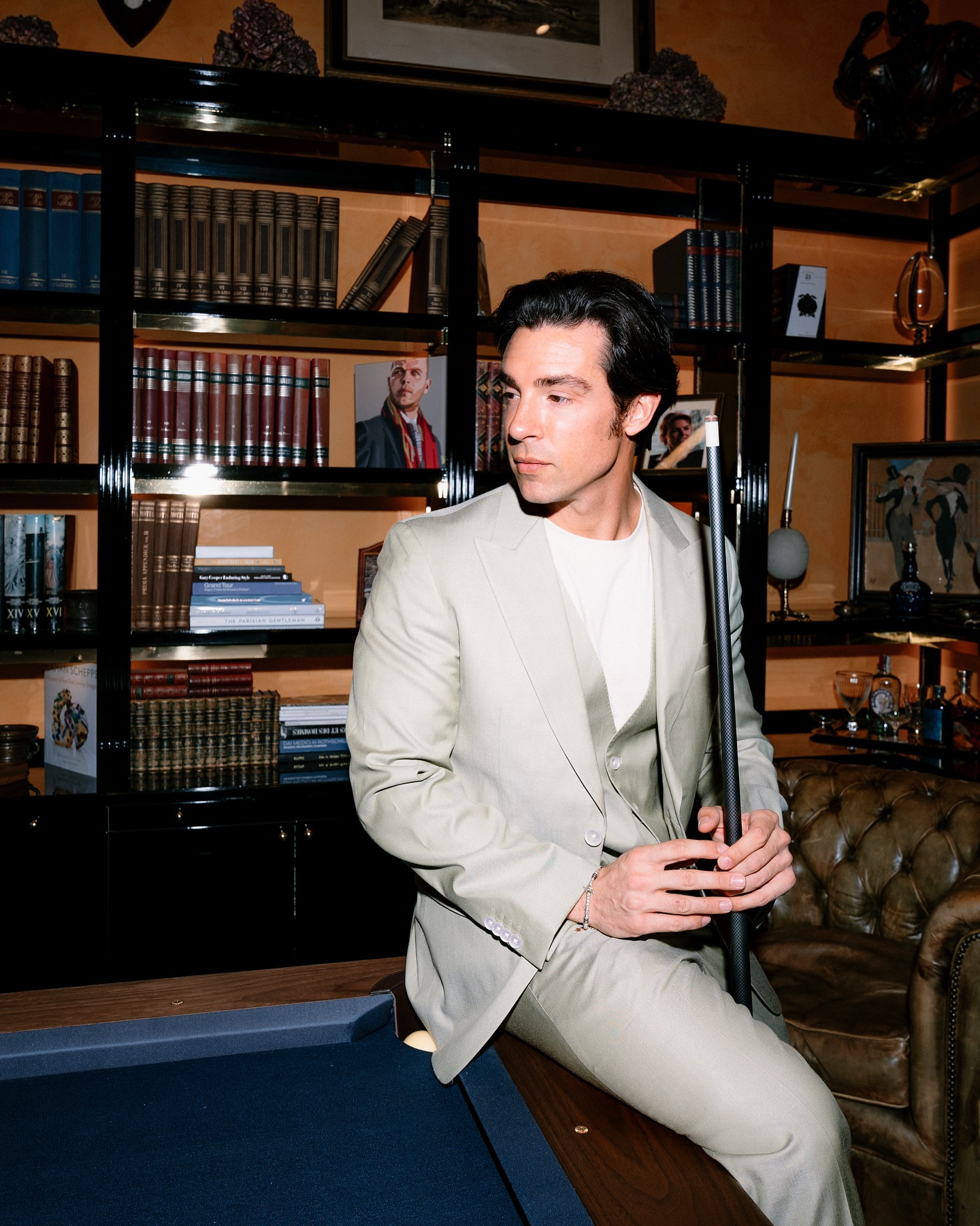 A man in a light beige suit sits on the edge of a pool table holding a cue stick, in a stylish library with shelves of books and art objects behind him.