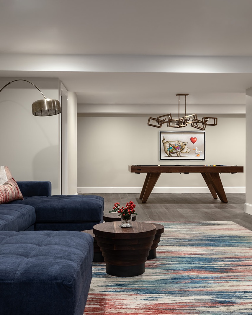 A modern living space with a blue sectional sofa, round wooden coffee table with red flowers, colorful striped rug, pool table, contemporary chandelier, and wall art in the background.