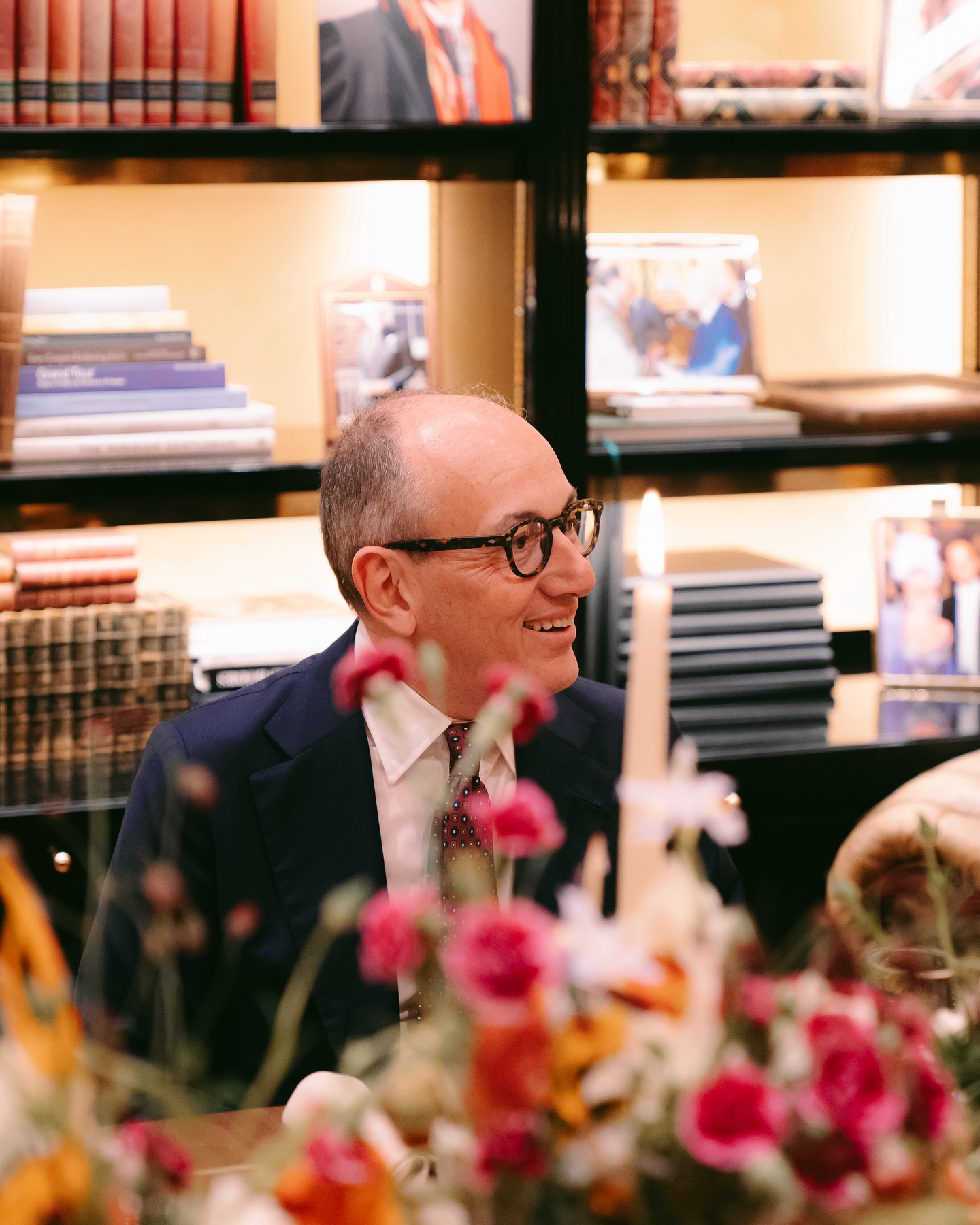 A man in glasses and a suit sits at a table with flowers and a lit candle, smiling. Behind him are shelves with books and framed photos. The scene is warm and inviting.