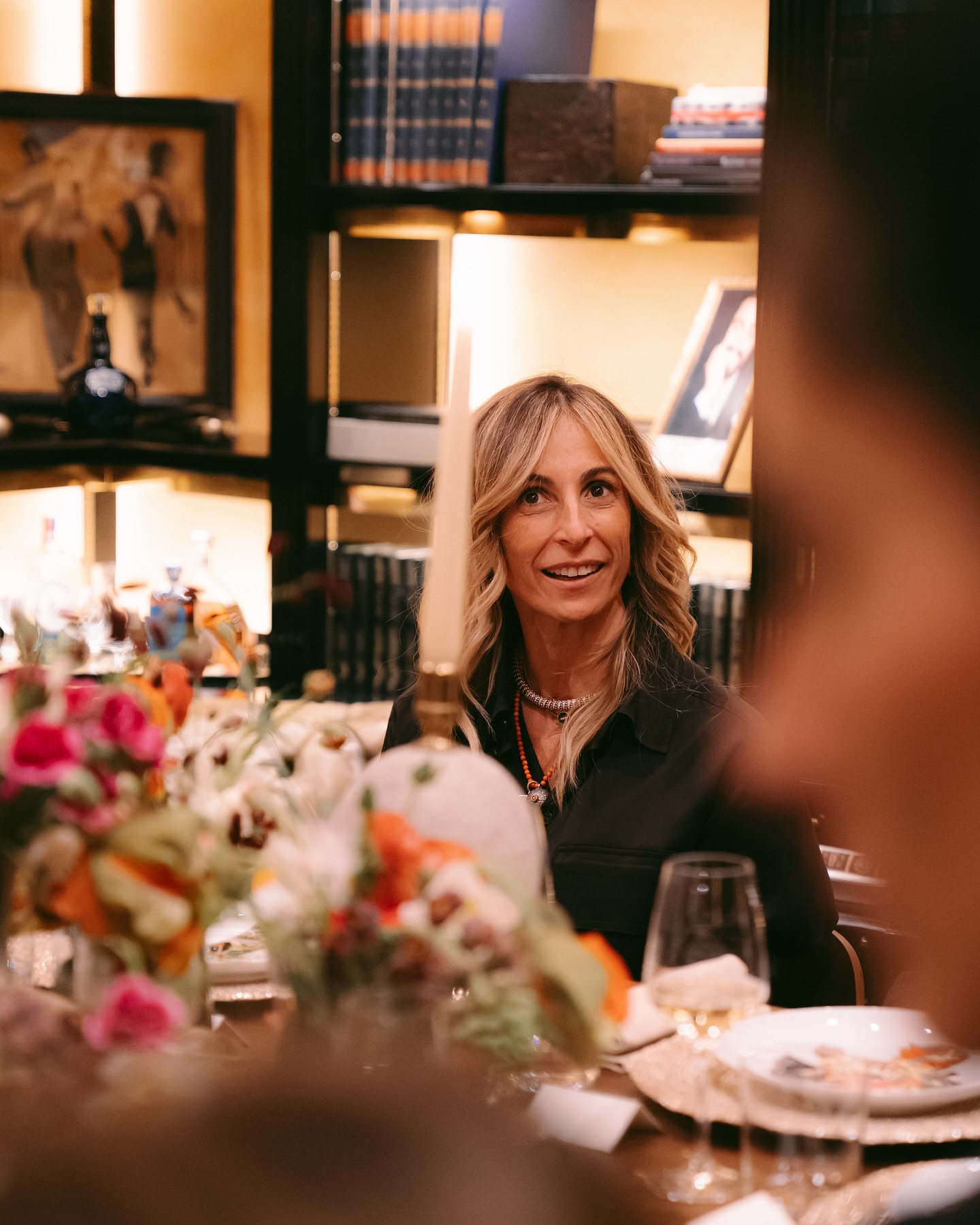A woman with wavy blonde hair sits at a dinner table decorated with colorful flowers, candles, and glassware, smiling. Shelves with books and framed art are visible in the warmly lit background.