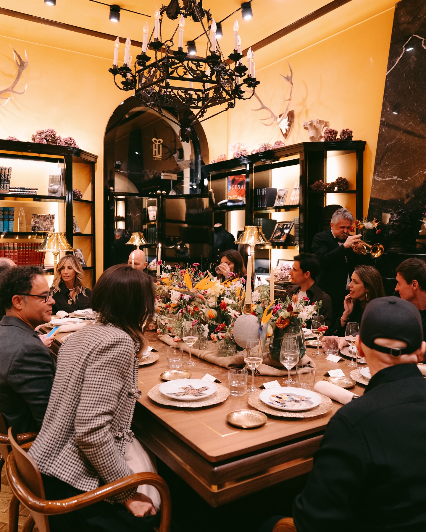 A group of people are seated around an elegantly set dining table in a warmly lit, upscale room, while a musician plays a trumpet in the background. The shelves are filled with books and decor, and a chandelier hangs above.