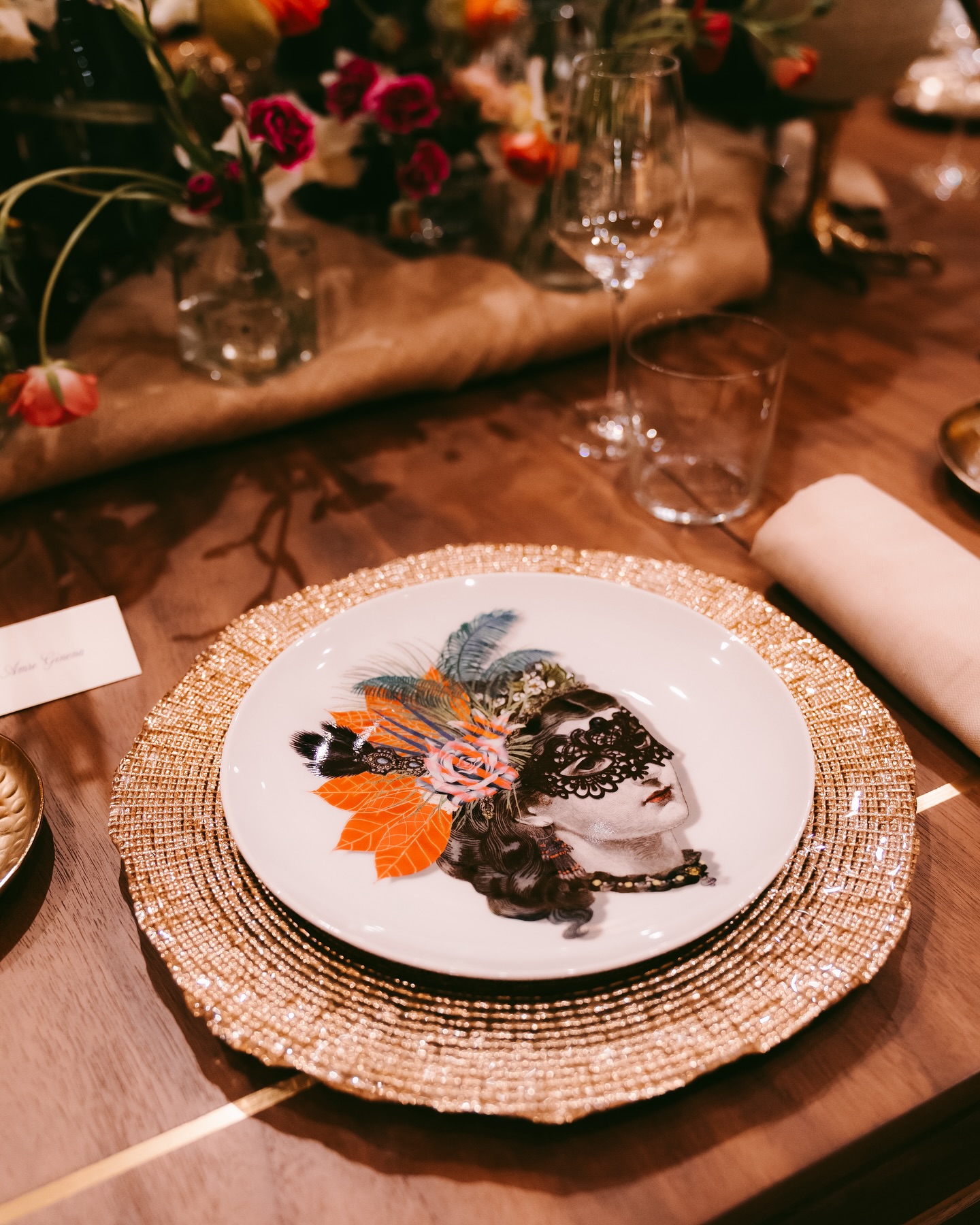 A decorative dinner plate with an artistic image of a womans masked face, adorned with colorful feathers and flowers, sits on a gold beaded charger at an elegantly set table with glassware, flowers, and a napkin.