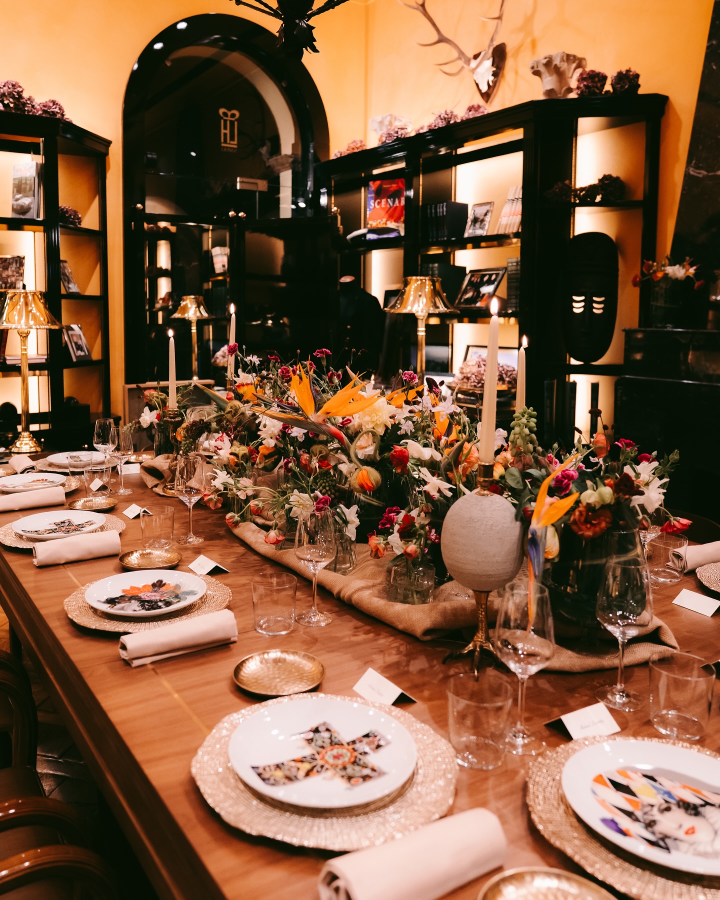A long, elegant dining table is set for a formal meal with floral centerpieces, candles, gold chargers, folded napkins, and glassware. Shelves with books and decor line the warmly lit, yellow-walled room.