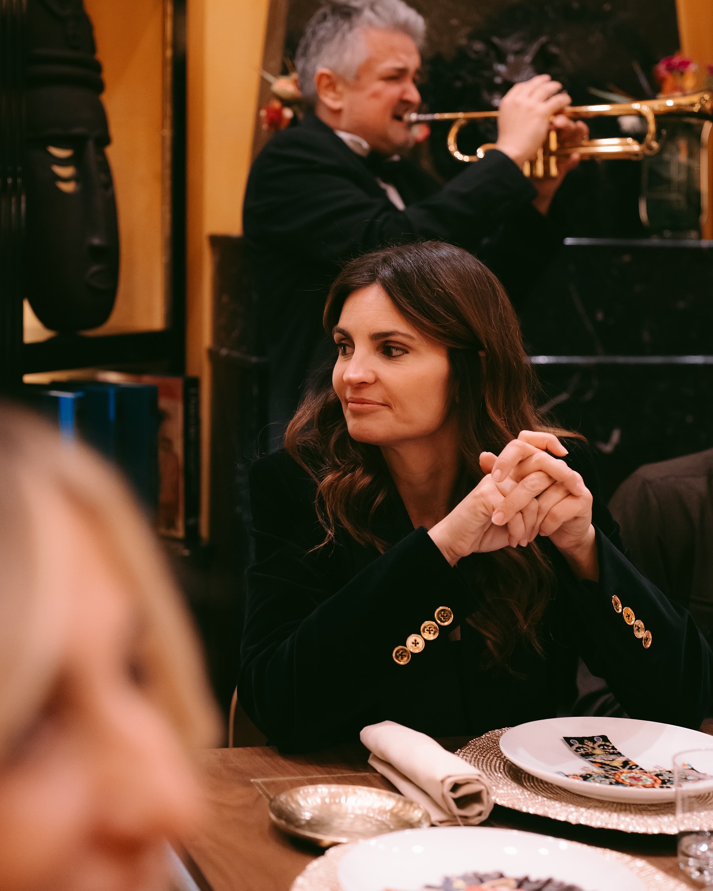 A woman with long brown hair sits at a dining table with her hands clasped, looking to the side. Behind her, a musician plays a trumpet. The table is set with plates, a napkin, and cutlery.