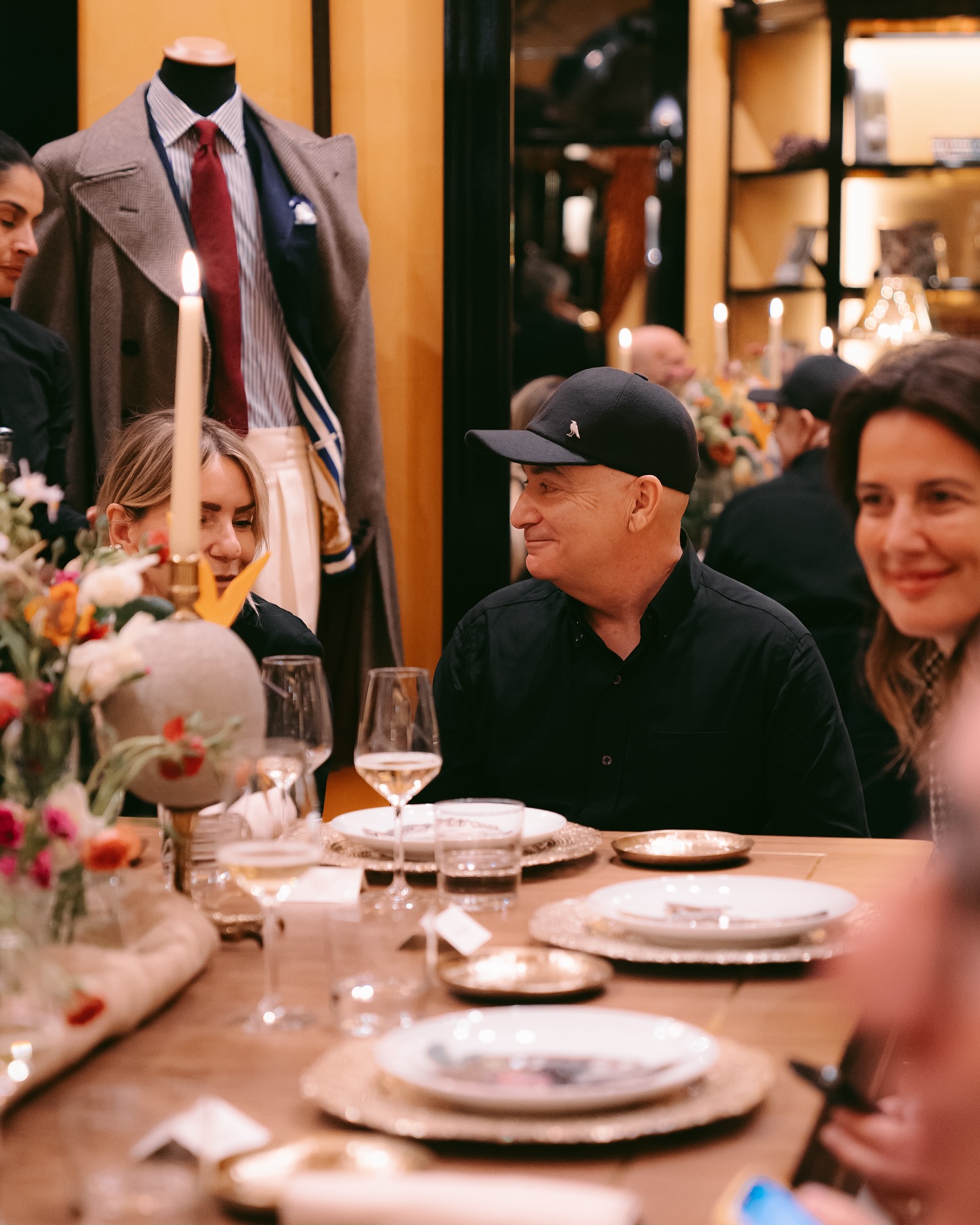 People sitting around a candlelit dinner table set with flowers, wine glasses, and plates; a man in a black shirt and cap smiles at a woman beside him. A mannequin dressed in a coat is visible in the background.
