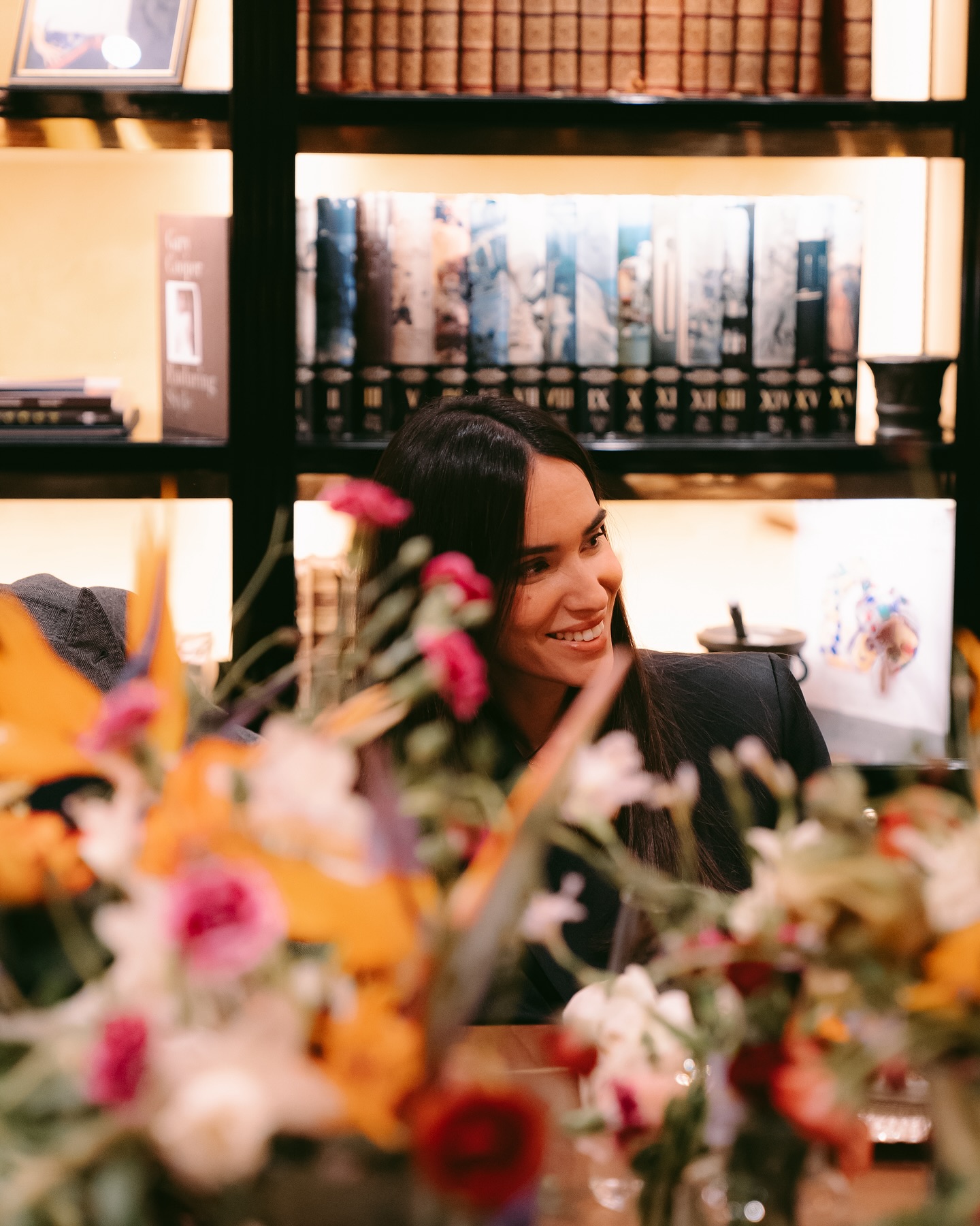 A woman with long dark hair smiles while sitting at a table decorated with colorful flowers, with bookshelves filled with books in the background.