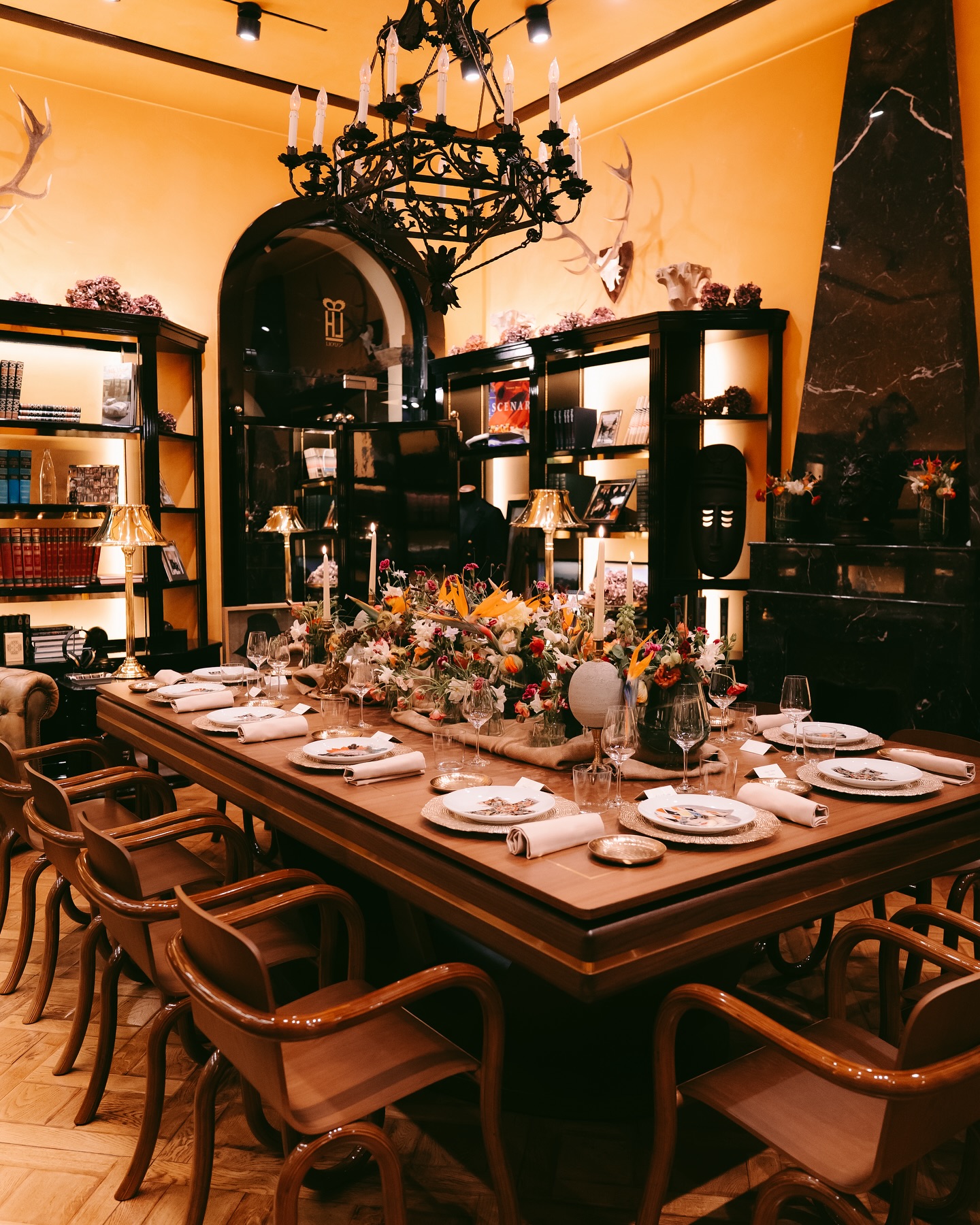 Elegant dining room with a large wooden table set for a meal, surrounded by chairs. The table has floral centerpieces and candles. Bookshelves line the walls, and a black chandelier hangs from the ceiling. The lighting is warm and inviting.