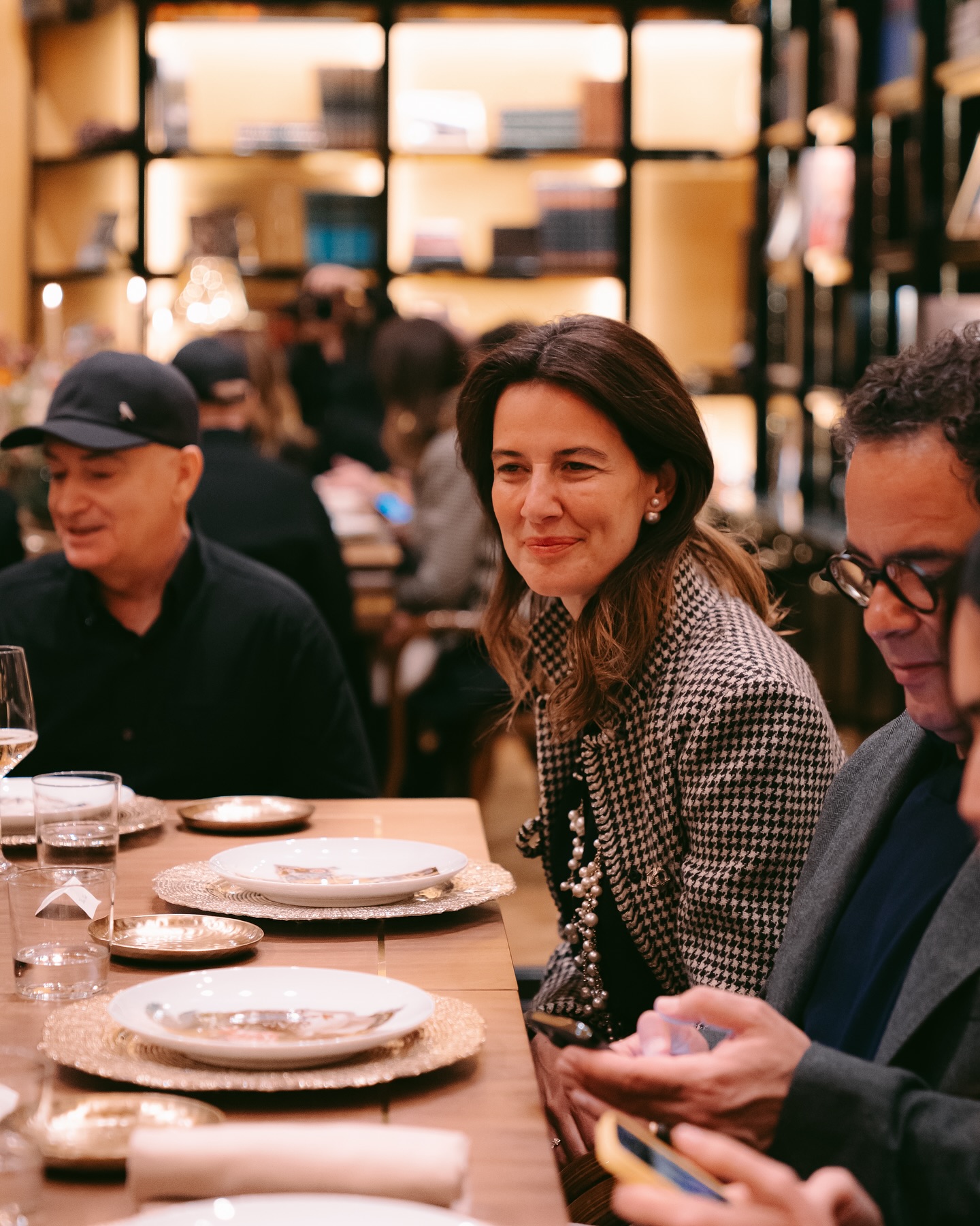 A woman in a checkered blazer smiles while seated at a dining table set with plates and glasses, surrounded by other people in a warmly lit, elegant restaurant.