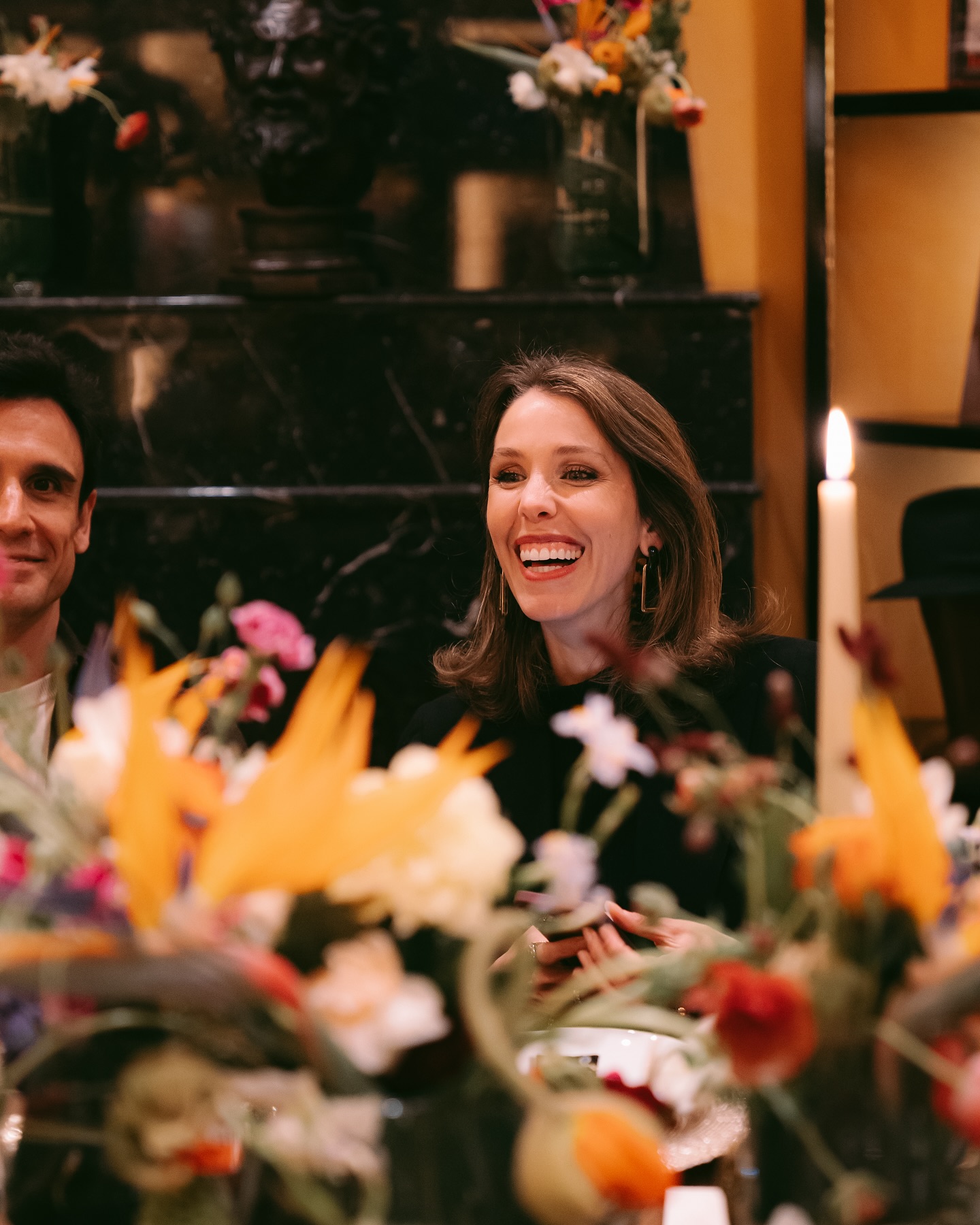 A woman with shoulder-length brown hair is smiling and laughing at a candle-lit dinner table, surrounded by colorful flowers and a man seated beside her.