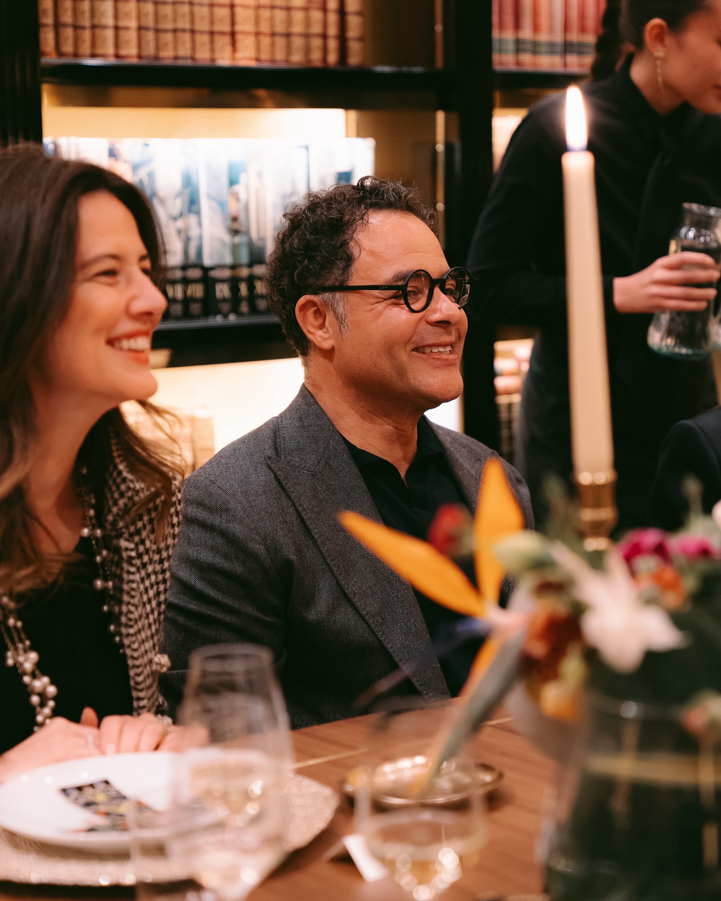 Two people sit smiling at a warmly lit dinner table with a tall candle, flowers, and bookshelves in the background. One person pours water in the background. The atmosphere appears elegant and joyful.