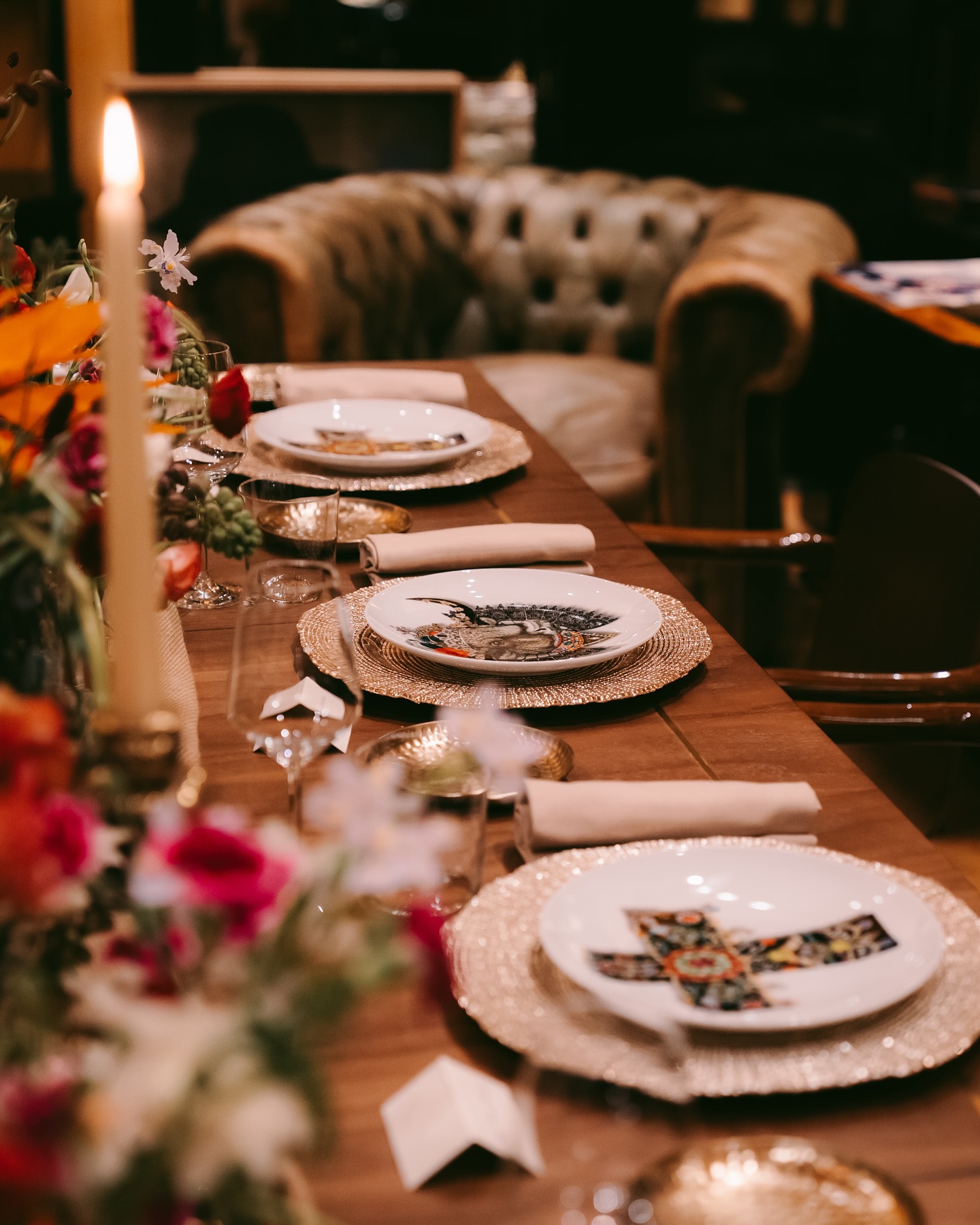 A wooden dining table set with decorative plates, woven placemats, napkins, wine glasses, candles, and colorful flowers, with a plush armchair in the background.