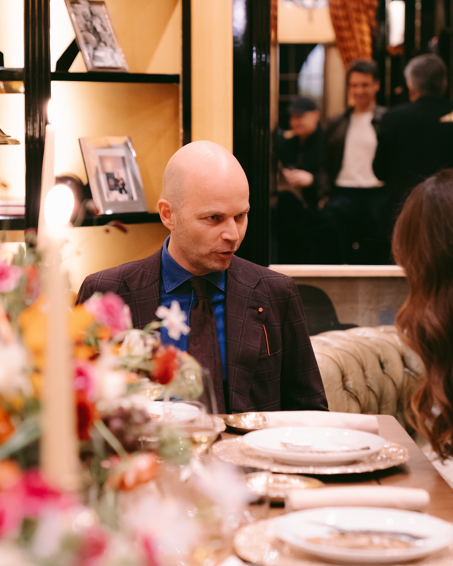 A bald man in a suit sits at a dining table set with plates, candles, and colorful flowers, facing a woman with wavy brown hair. People are visible in the background, reflected in a mirror.