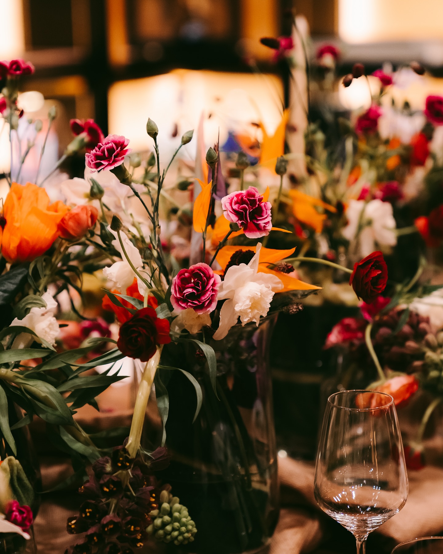 A close-up of a table decorated with vibrant flower arrangements in various colors, including red, pink, orange, and white, with an empty wine glass in the foreground.